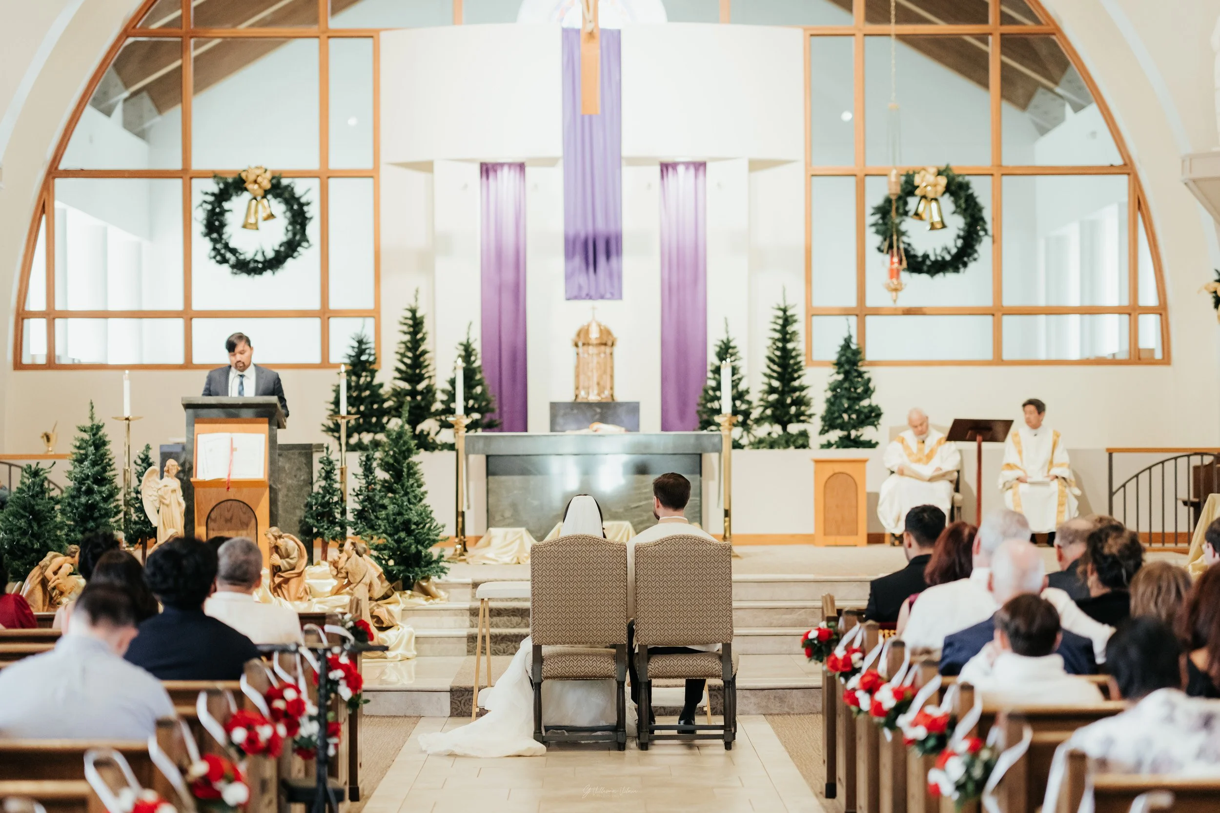 A wedding ceremony inside a church decorated with Christmas wreaths, ornaments, and small pine trees, with a bride and groom sitting at the altar facing the officiant.