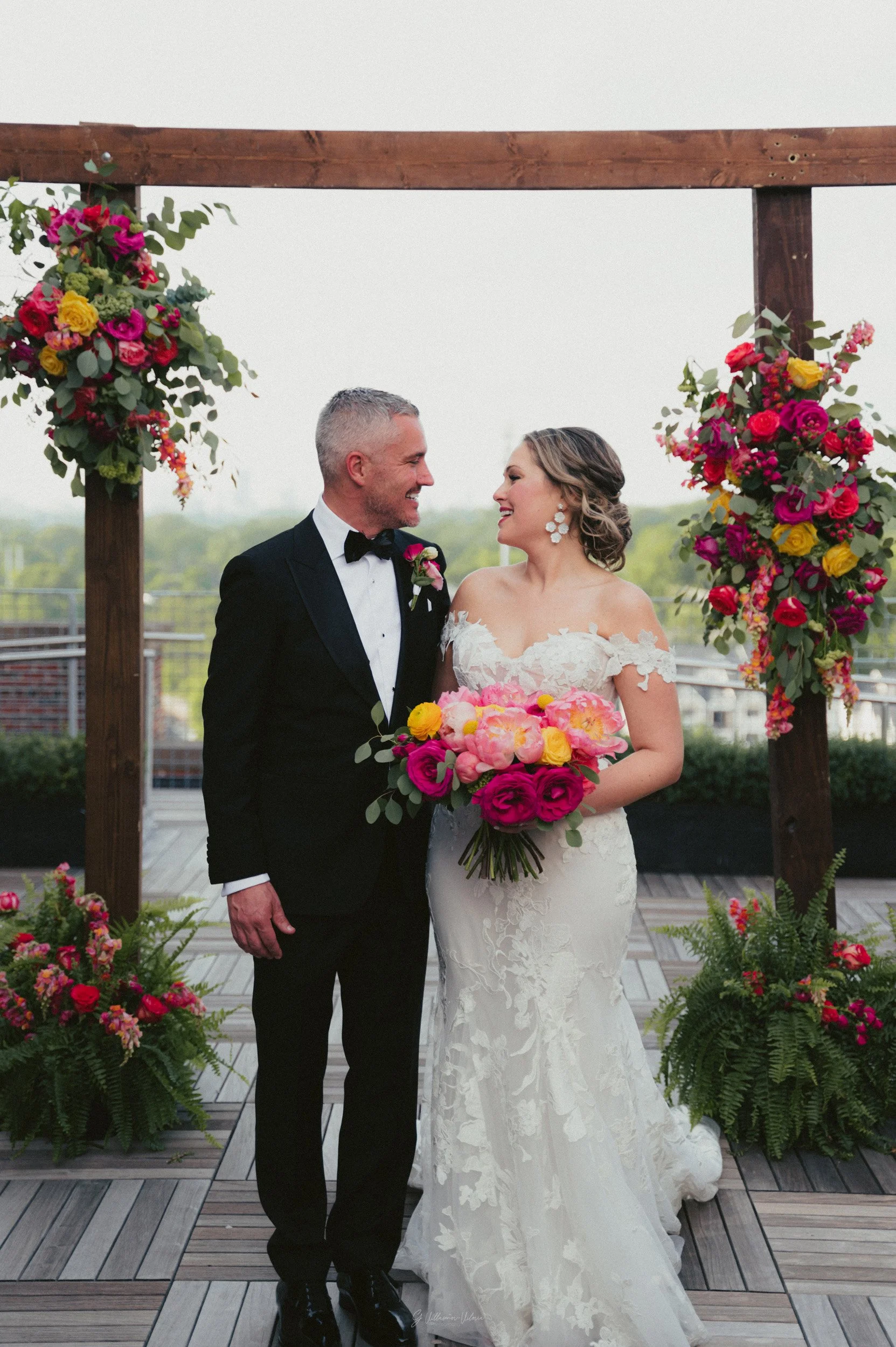 A bride and groom are standing together at their wedding ceremony, smiling at each other. The bride is wearing a white lace wedding gown and holding a bouquet of pink, yellow, and green flowers. The groom is dressed in a black tuxedo with a bow tie. 