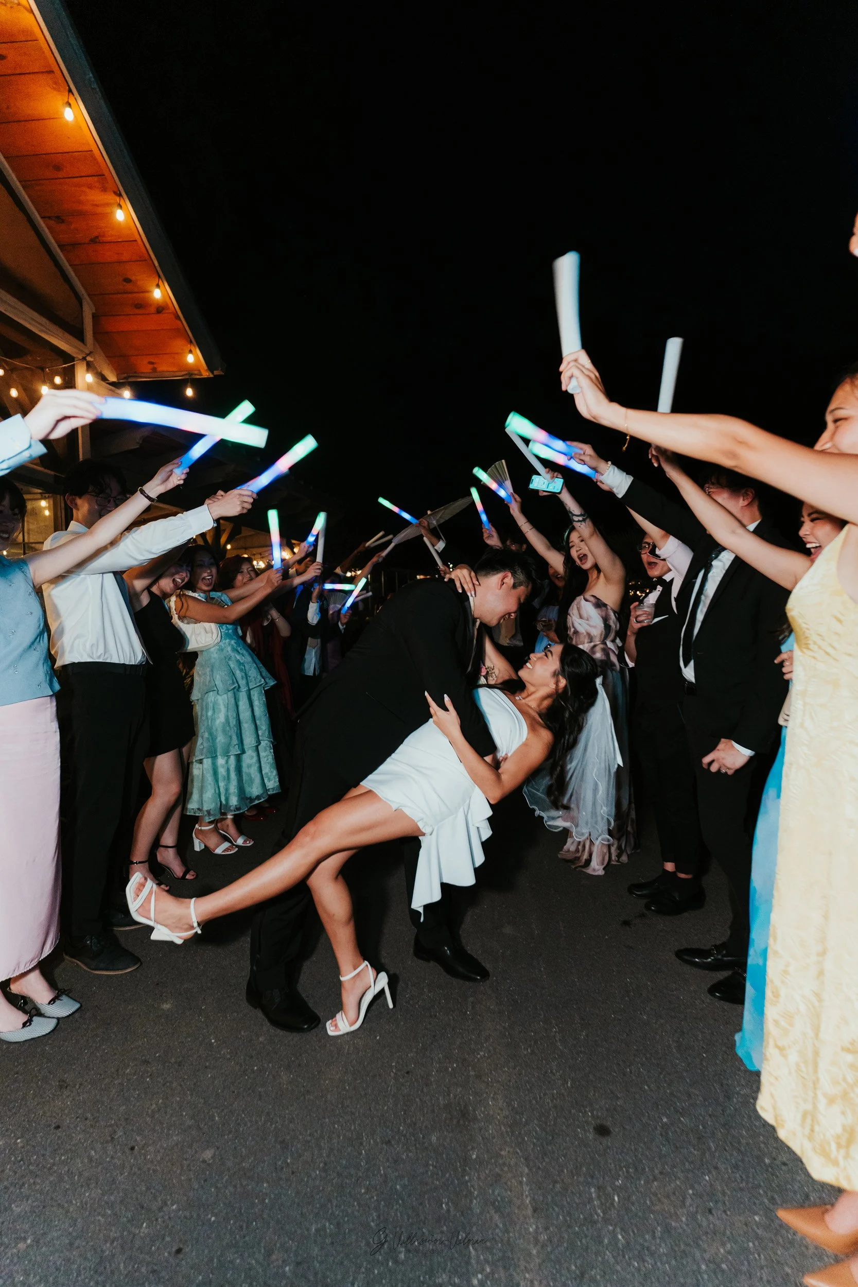 A newlywed couple dancing and kissing at their wedding reception while friends celebrate with glow sticks around them at night.