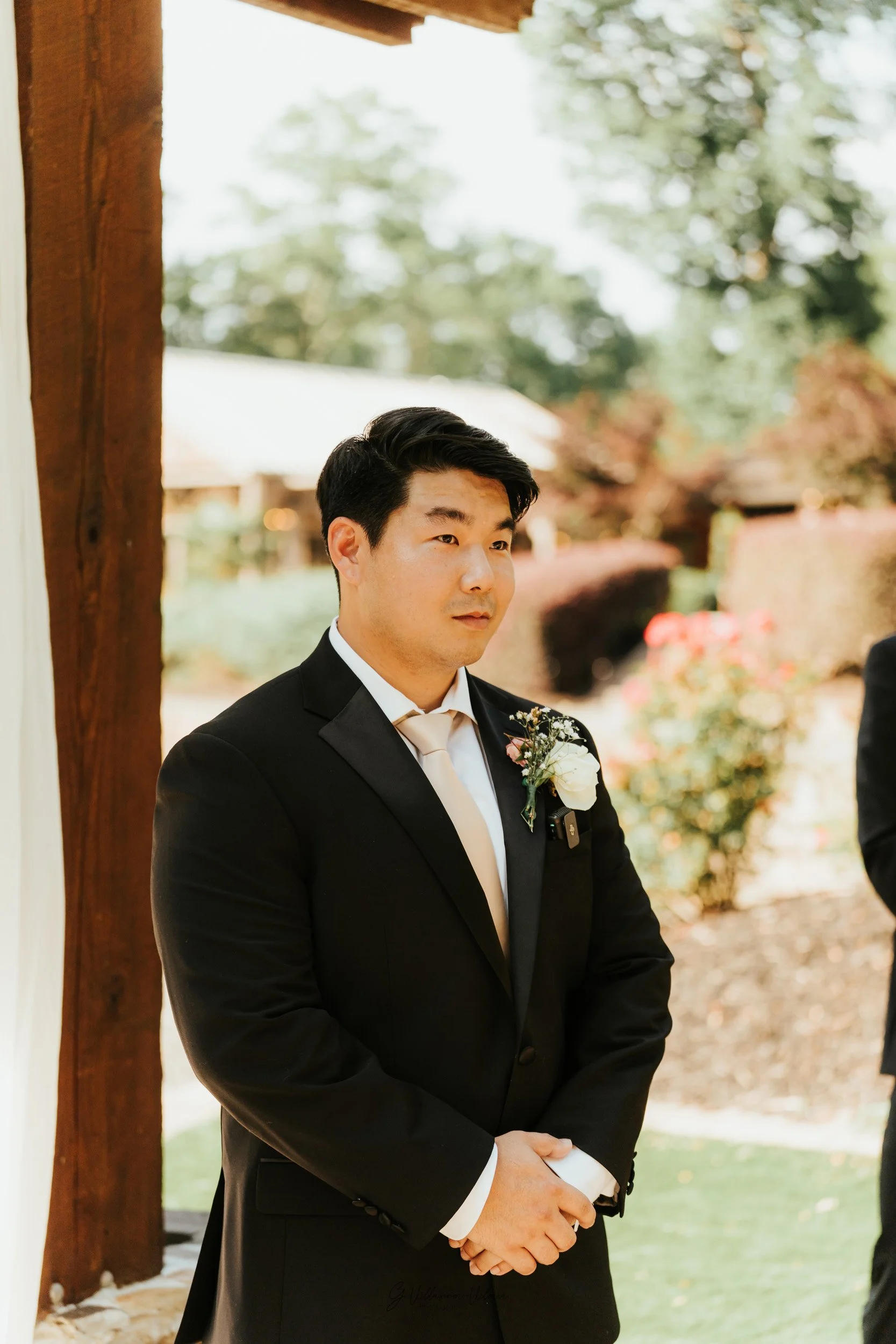 A man in a black tuxedo with a boutonniere stands outdoors at a wedding ceremony.