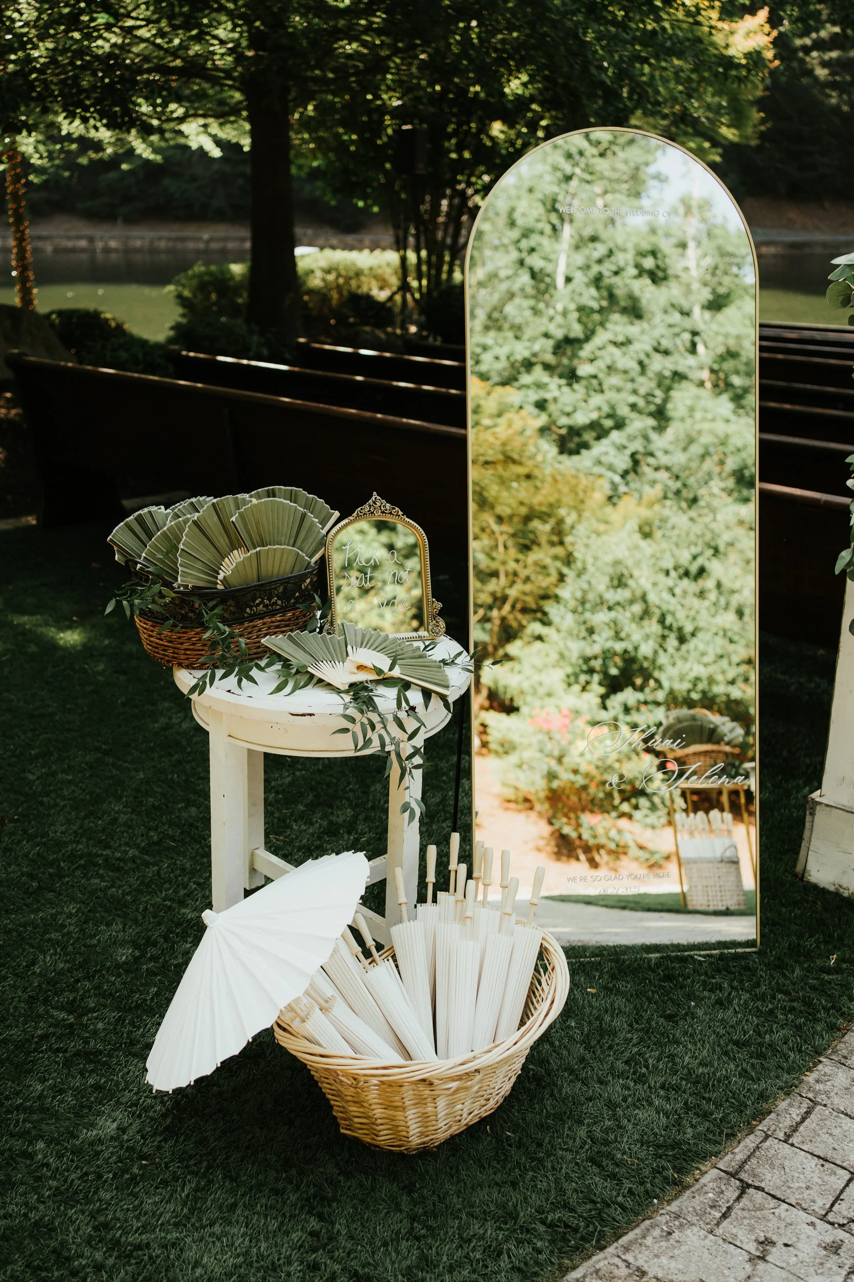 Outdoor wedding decor with a large, arched mirror reflecting greenery and a basket of paper fans, a white parasol, and a small table with a decorative mirror and greenery, set on a grassy area near water.