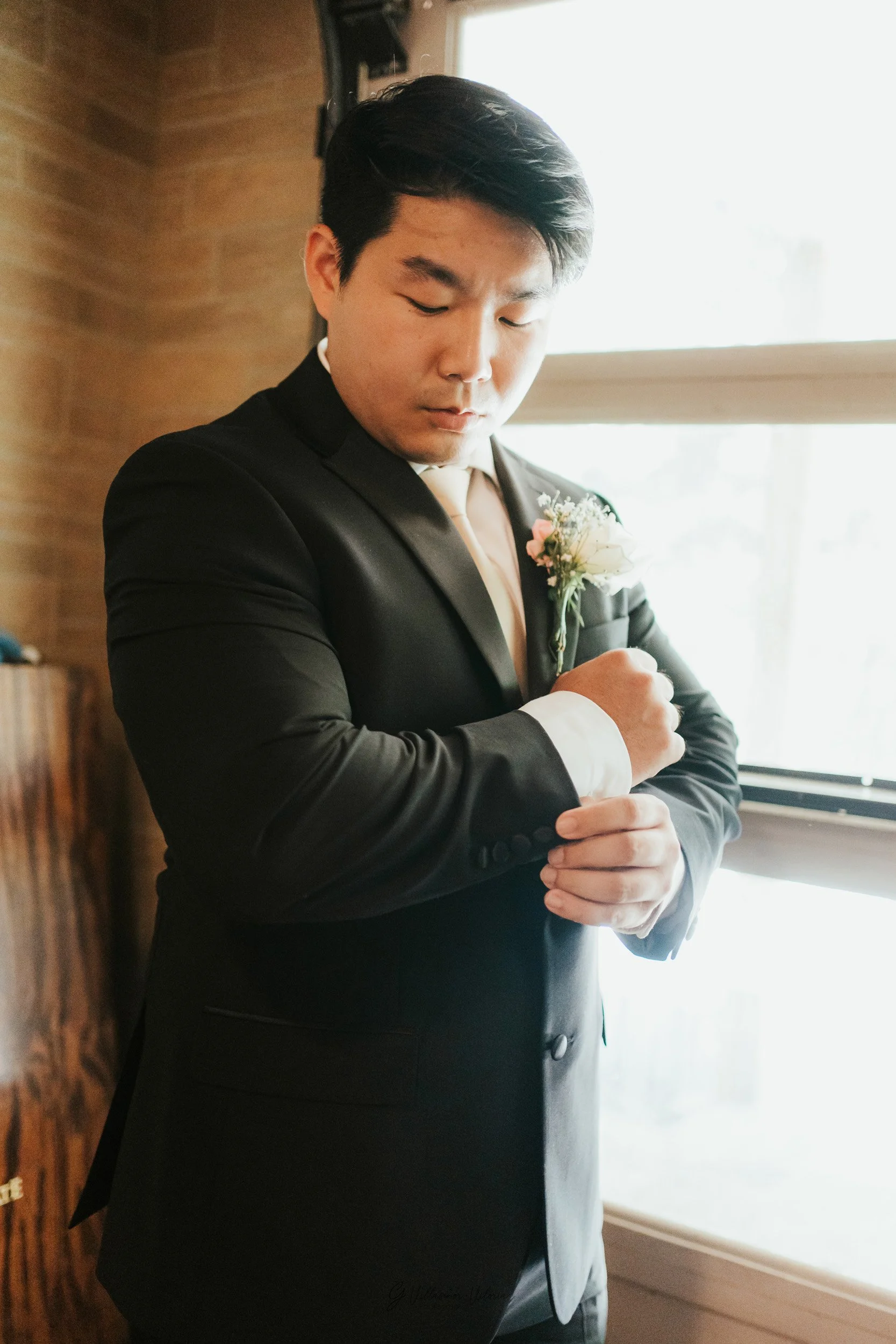 A man in a black suit with a boutonniere adjusts his cufflinks near a window with sunlight brightening the room.
