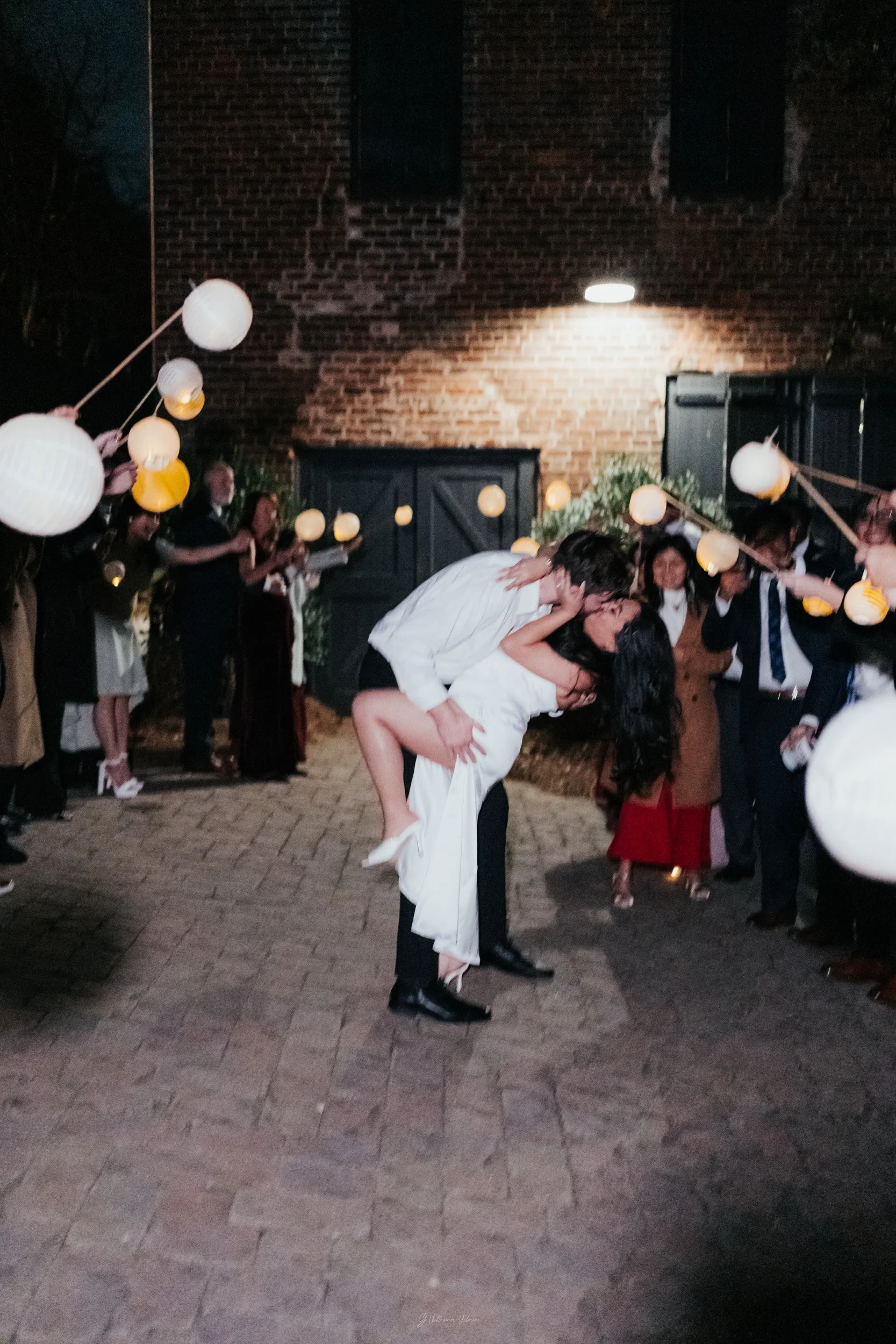 A couple at a wedding celebration dancing and kissing, with guests watching and balloons hanging overhead, outdoors at night against a brick wall.