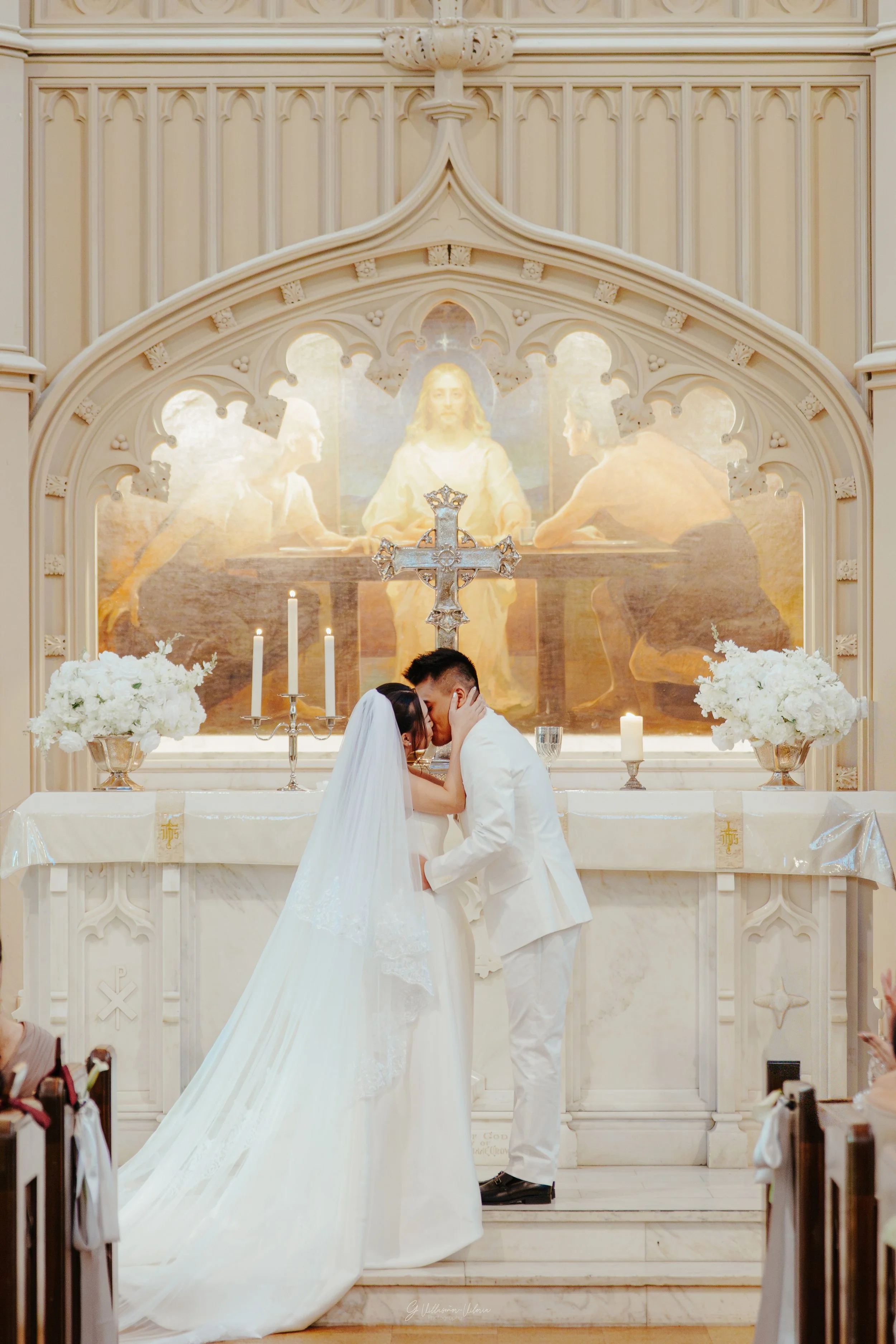 A bride and groom share a kiss at their wedding ceremony in a church, with a religious altar and a painting of Jesus Christ in the background.