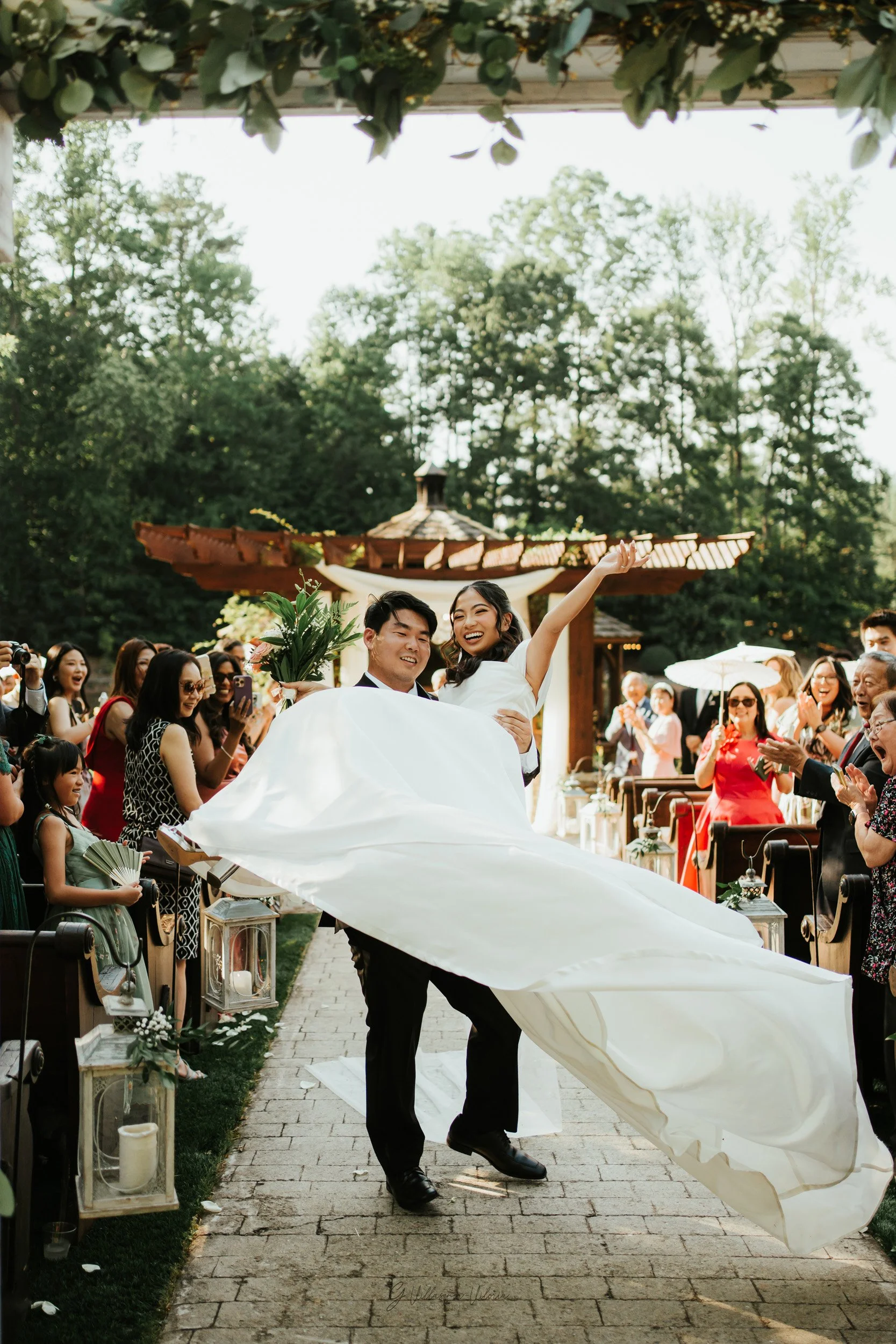 A newlywed couple celebrating their wedding outdoors, with the groom carrying the bride who is smiling and raising her arm. Family and friends surround them, clapping and taking photos on a sunny day.