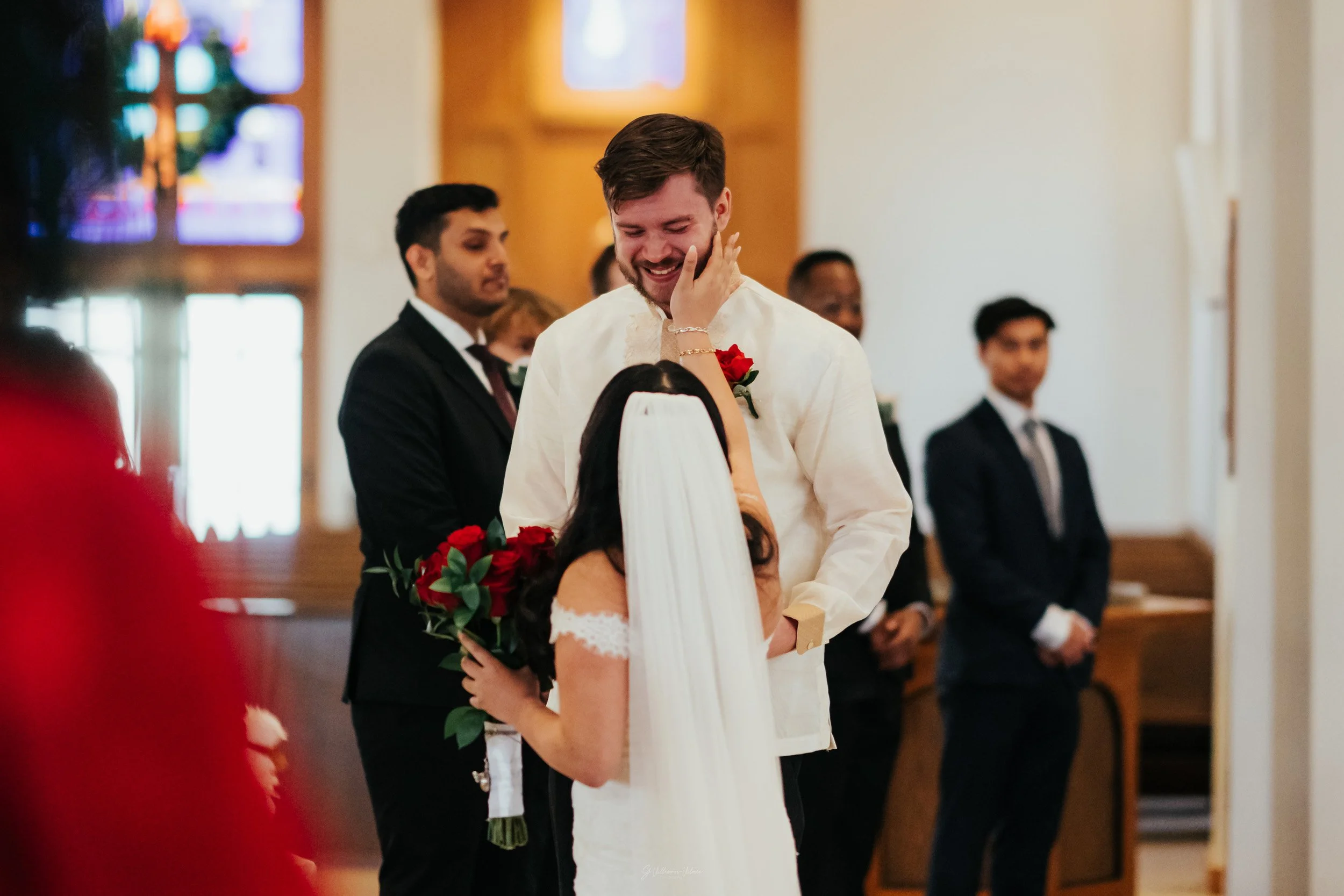 A bride and groom during their wedding ceremony, with the bride holding a bouquet of red roses and touching the groom's face who is smiling. There are witnesses or guests in suits standing behind them.