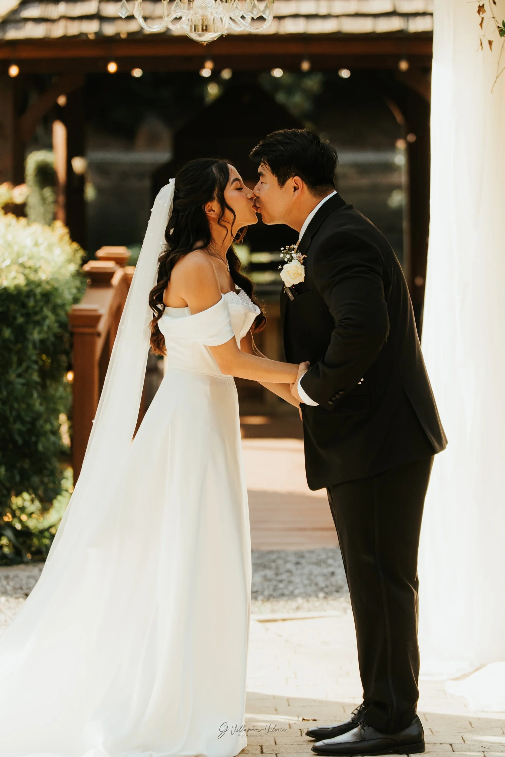 Bride and groom kissing during their wedding ceremony outdoors with greenery and a rustic backdrop.