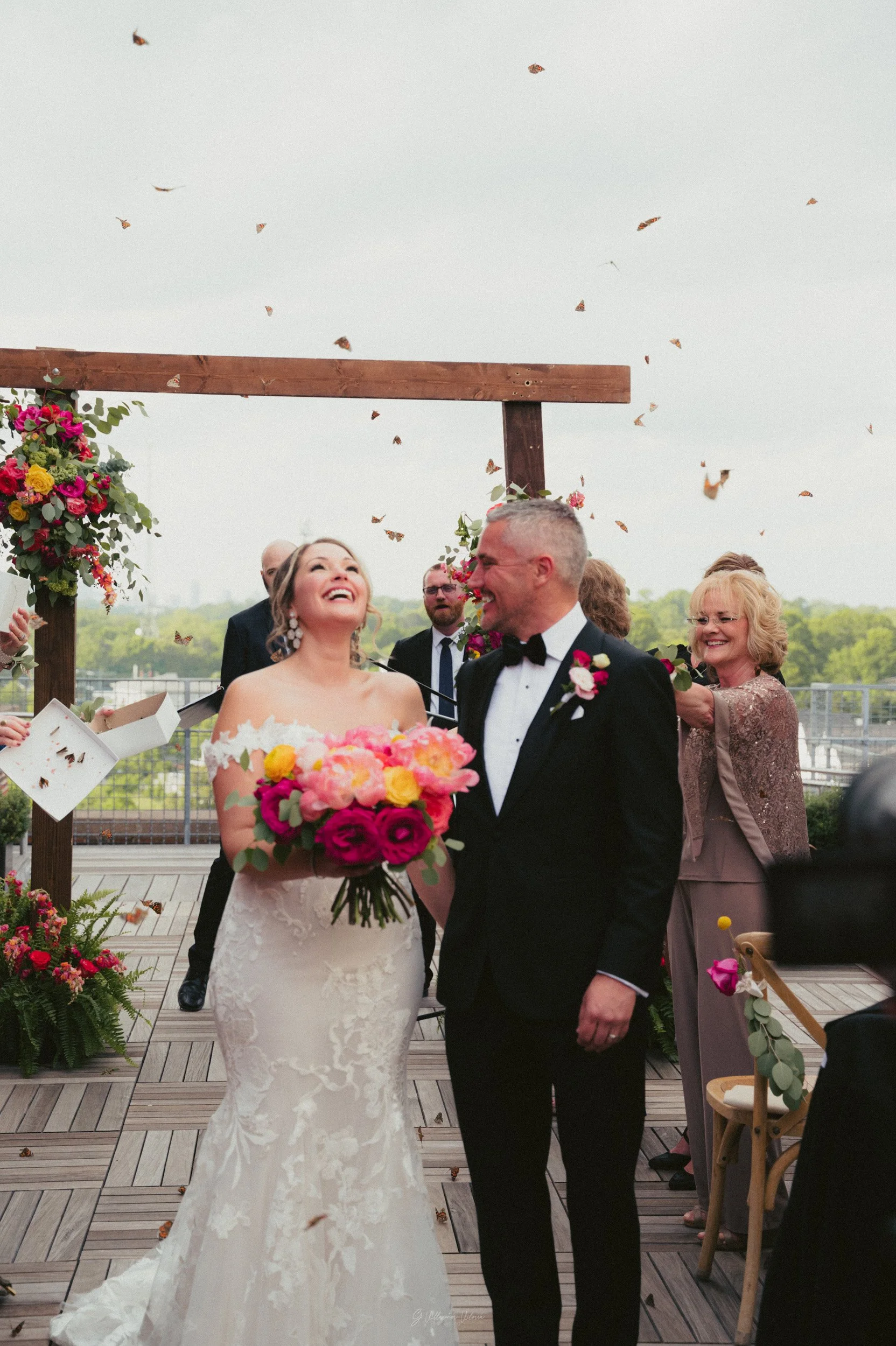 A bride and groom stand together smiling at their wedding ceremony, surrounded by friends and family, with butterflies flying above.