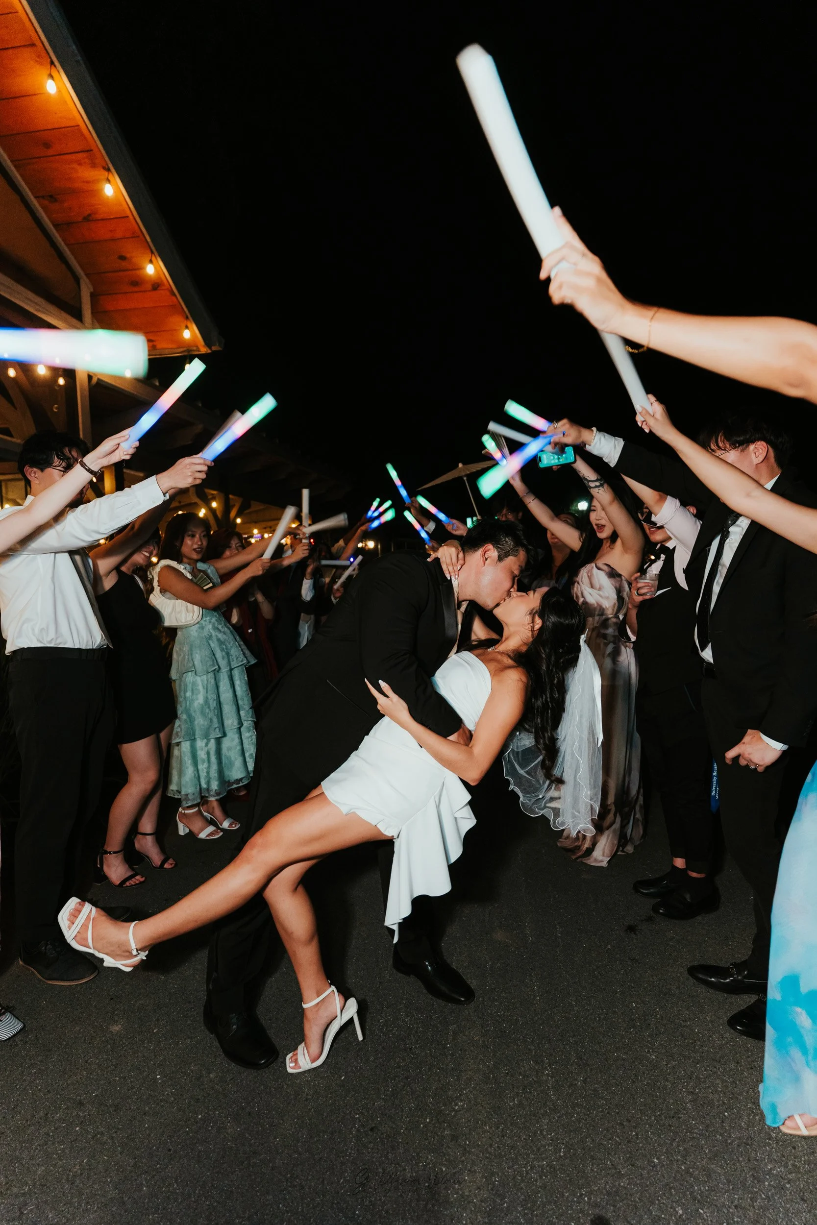 A newlywed couple sharing a romantic kiss at their wedding reception, surrounded by guests holding glow sticks and celebrating under string lights at night.