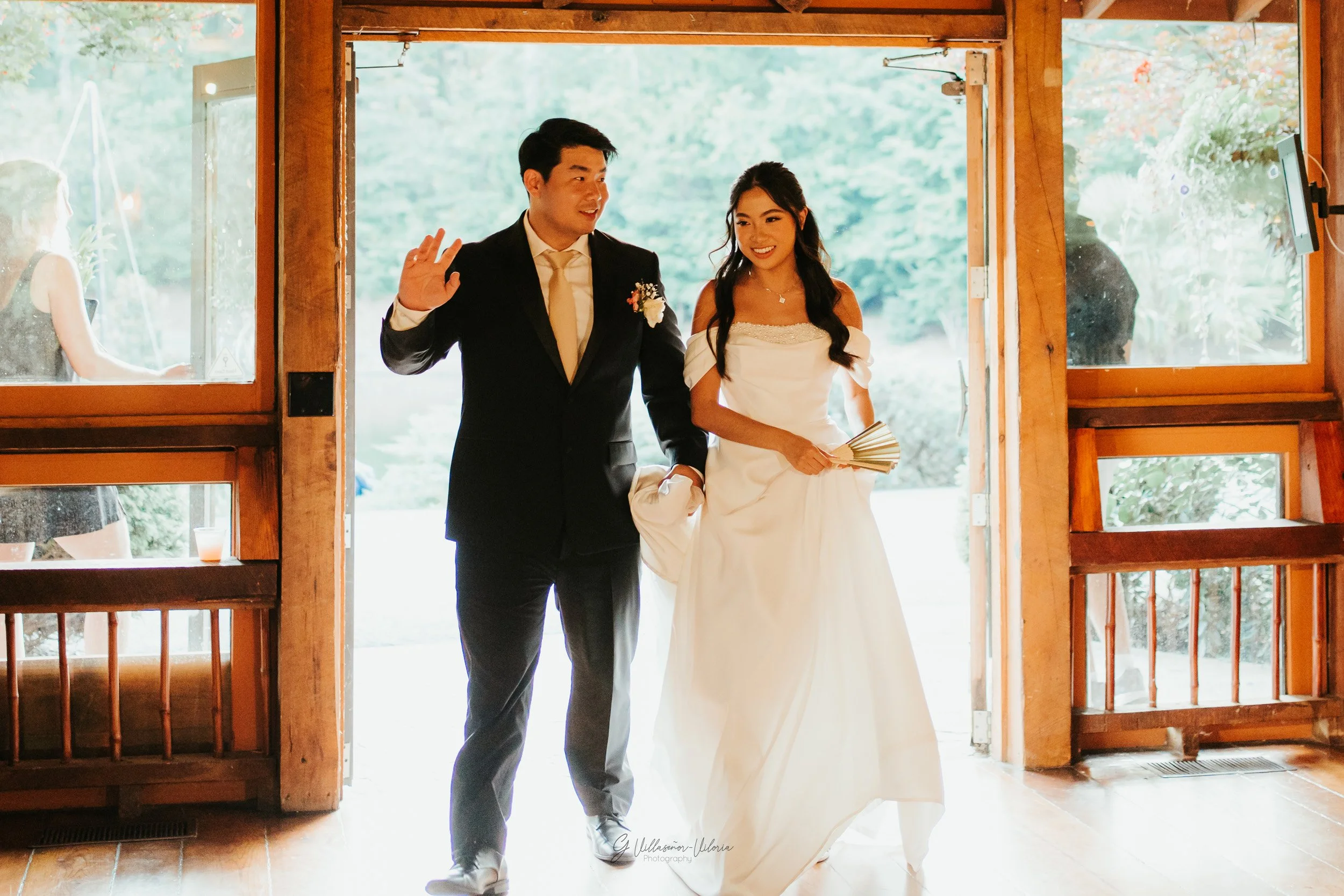 A bride and groom entering a venue, smiling, with the groom waving, inside a wooden building with open doors and greenery outside.