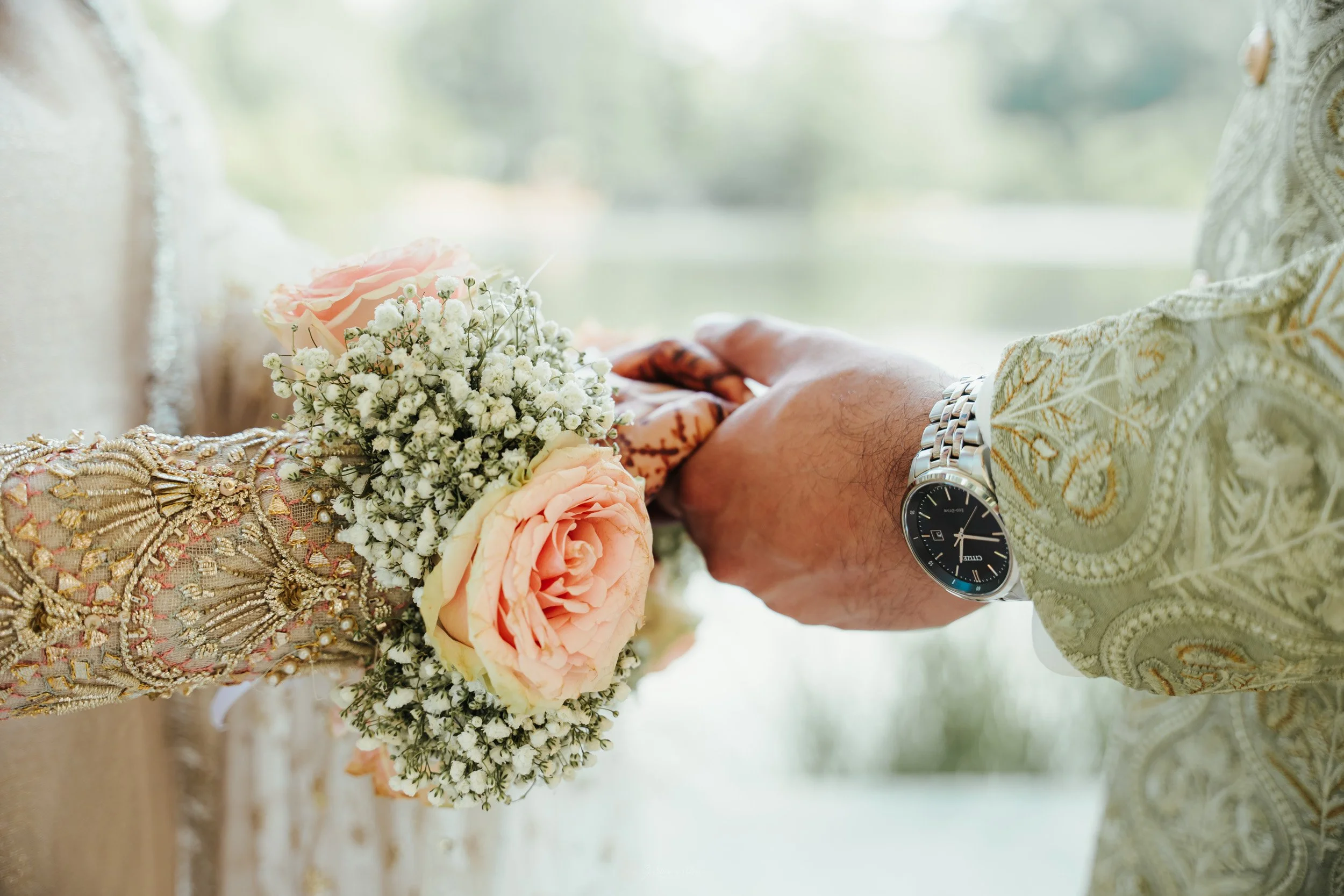 Close-up of a couple holding hands during a wedding, with the bride's hand holding a bouquet of pink roses and white baby's breath, and the groom wearing a patterned shirt with a silver watch.