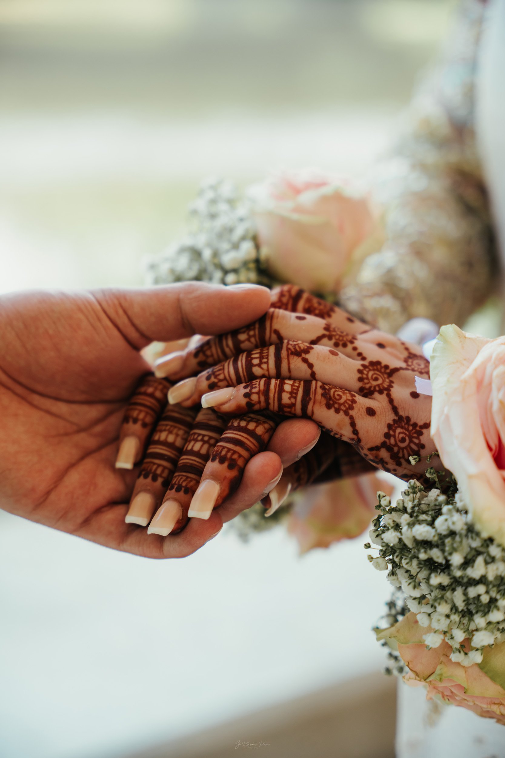 Close-up of two hands holding each other, one with intricate henna designs and long nails, surrounded by flowers including roses and baby's breath.