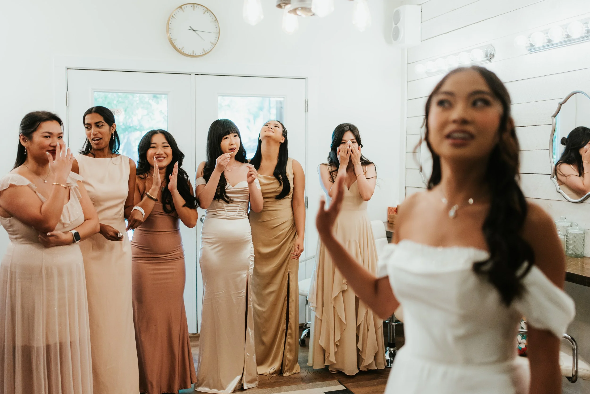 Bride in a white off-shoulder gown with dark wavy hair, gesturing with her hand, in front of a group of seven women in elegant dresses, reacting with joy and surprise inside a room with white walls, a clock, and a mirror.