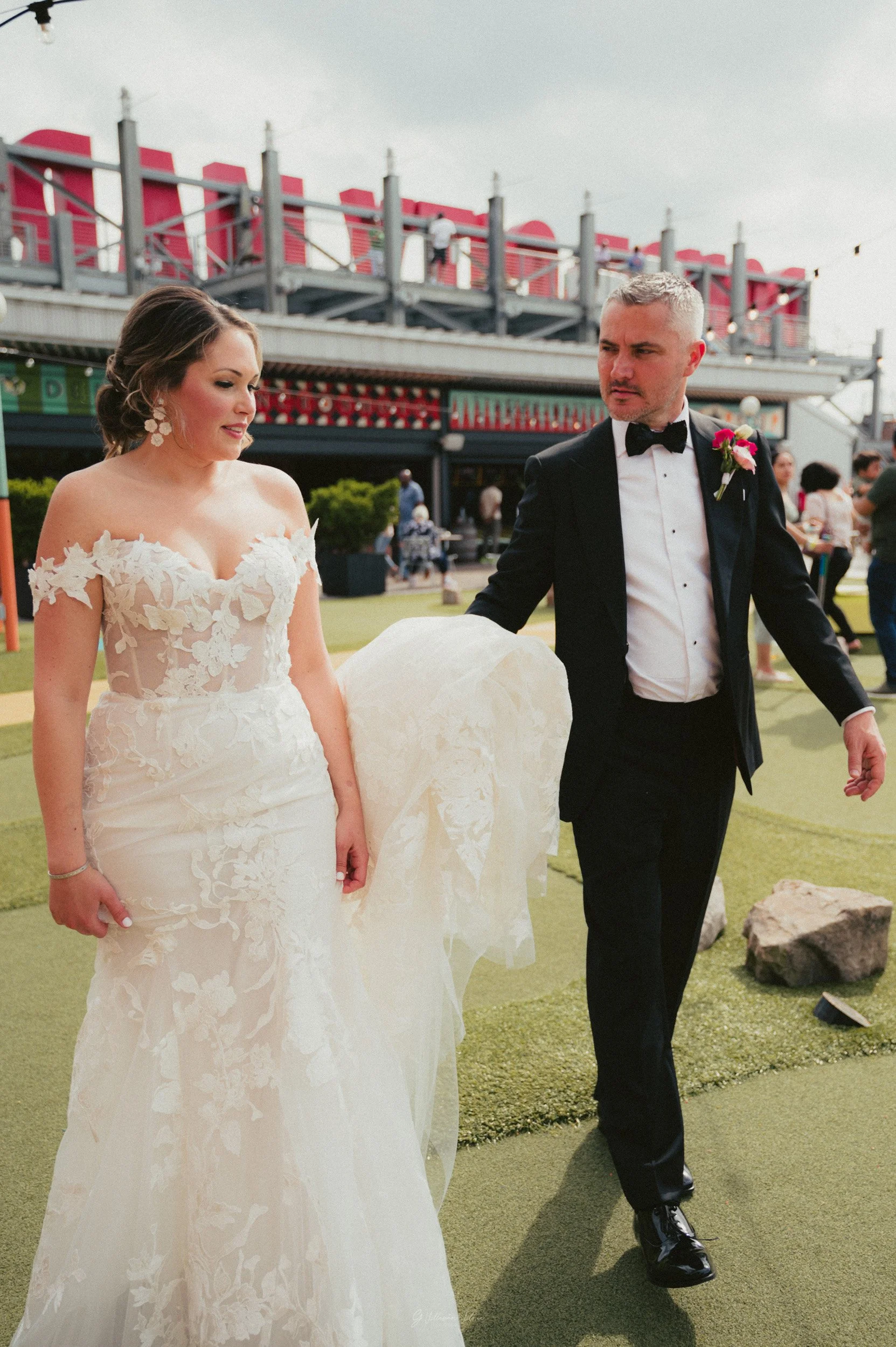 A bride and groom walking outside on a wedding day. The bride is wearing an off-the-shoulder white lace wedding gown and the groom is dressed in a black tuxedo with a bow tie and a boutonniere. They are on green artificial grass with rocks nearby, wi