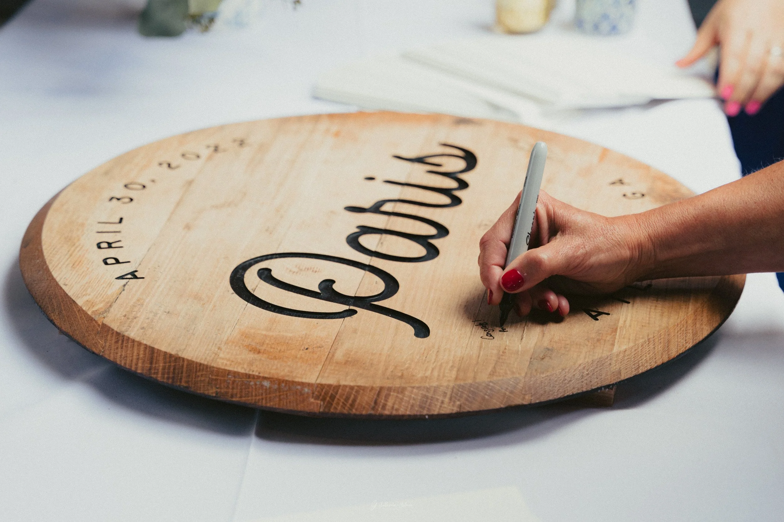 A person with red-painted nails signing a wooden wedding sign that reads 'April 30, 2023, Love'.
