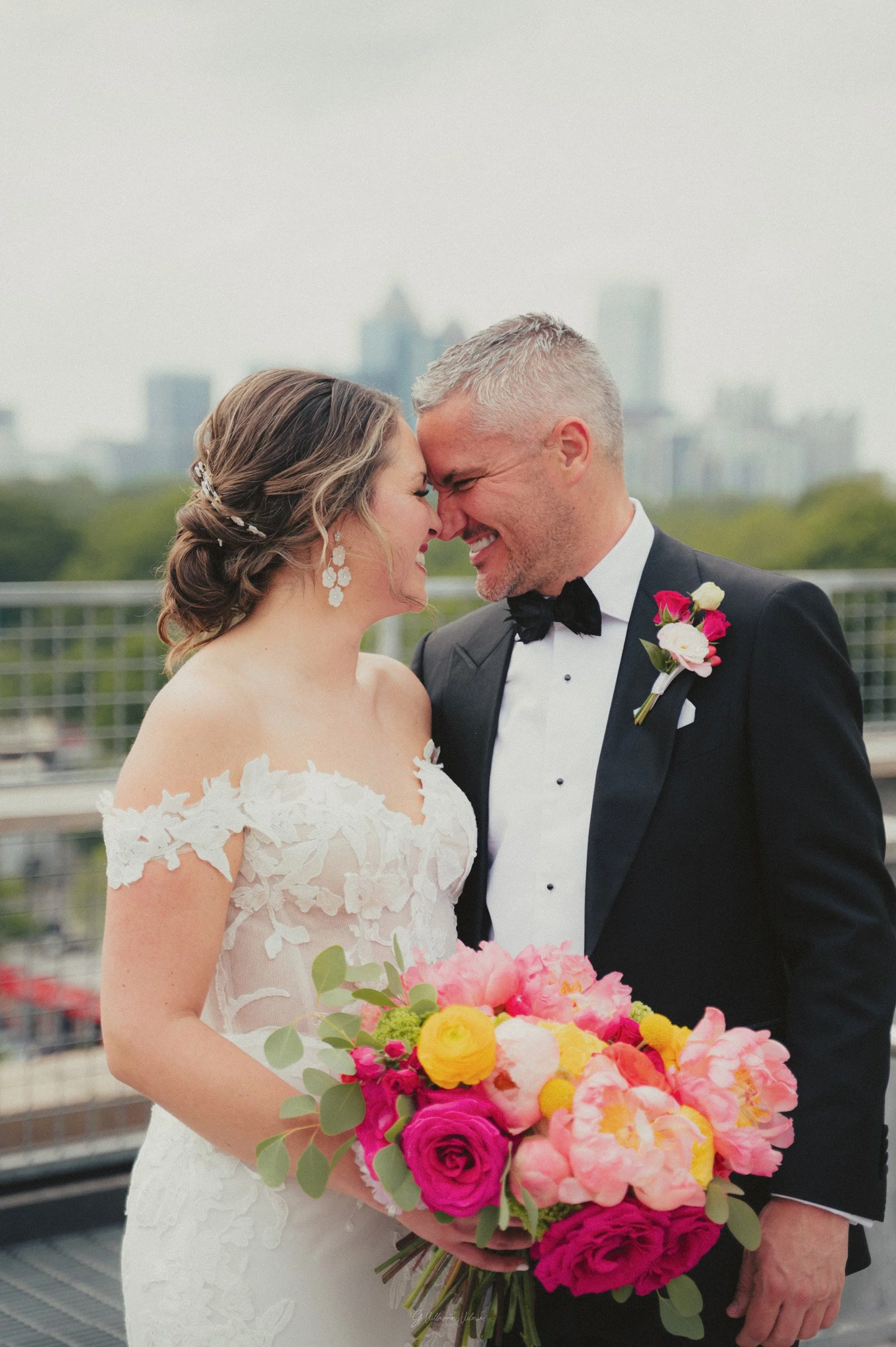 A bride and groom on their wedding day, touching foreheads and smiling, with a cityscape background, the bride holding a large bouquet of pink, yellow, and white flowers.
