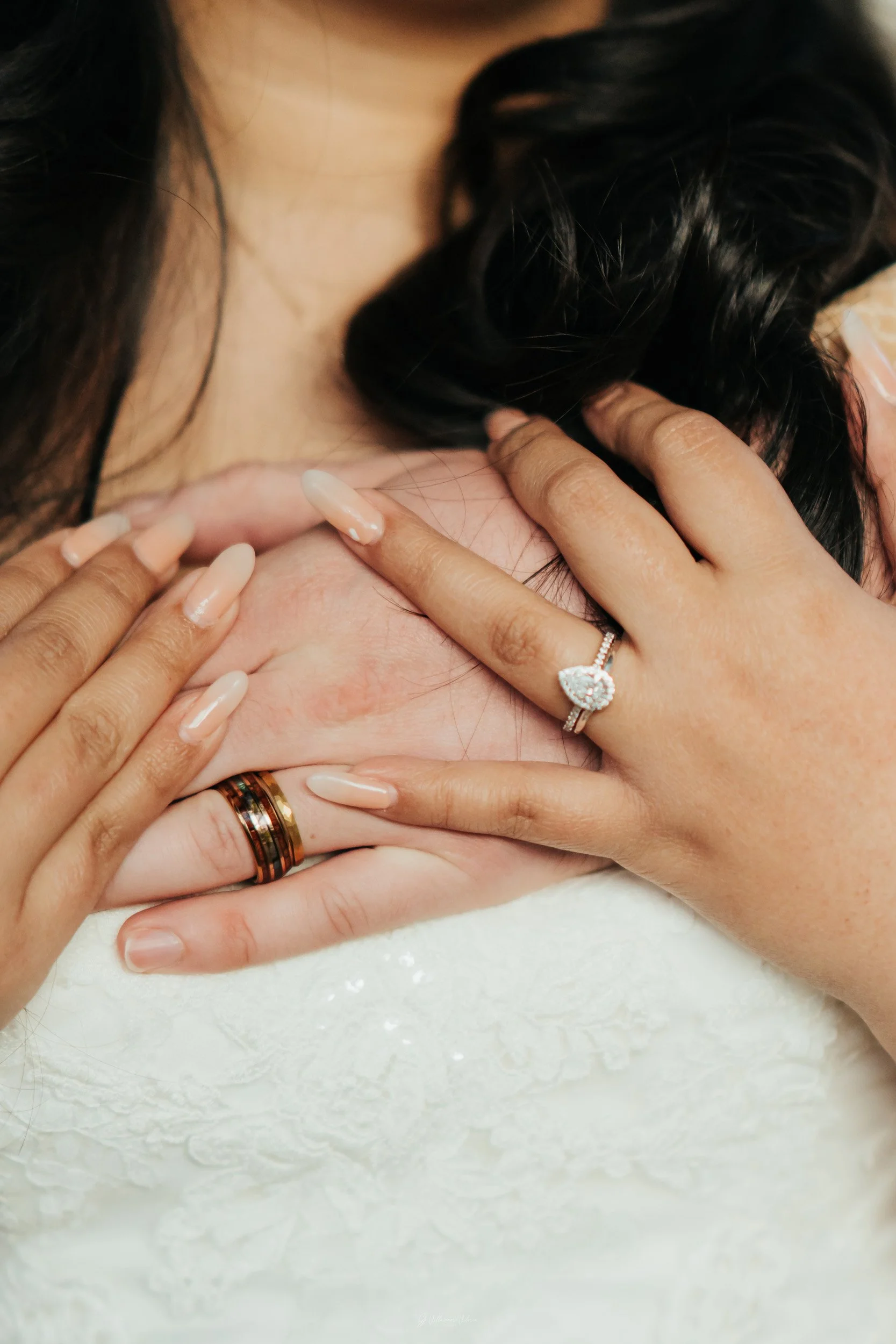 Close-up of two women touching each other's hands and neck, with one wearing a wedding ring and the other wearing a multicolored ring, against a white lace fabric background.