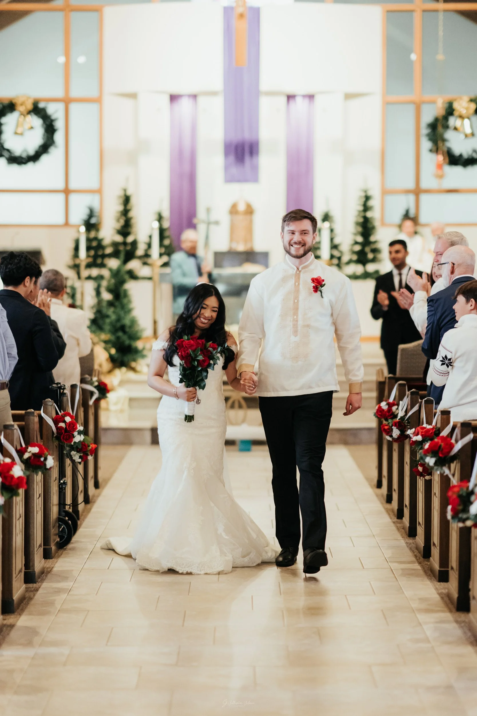 A bride and groom walking down the aisle in a church decorated for Christmas, with guests clapping and smiling.