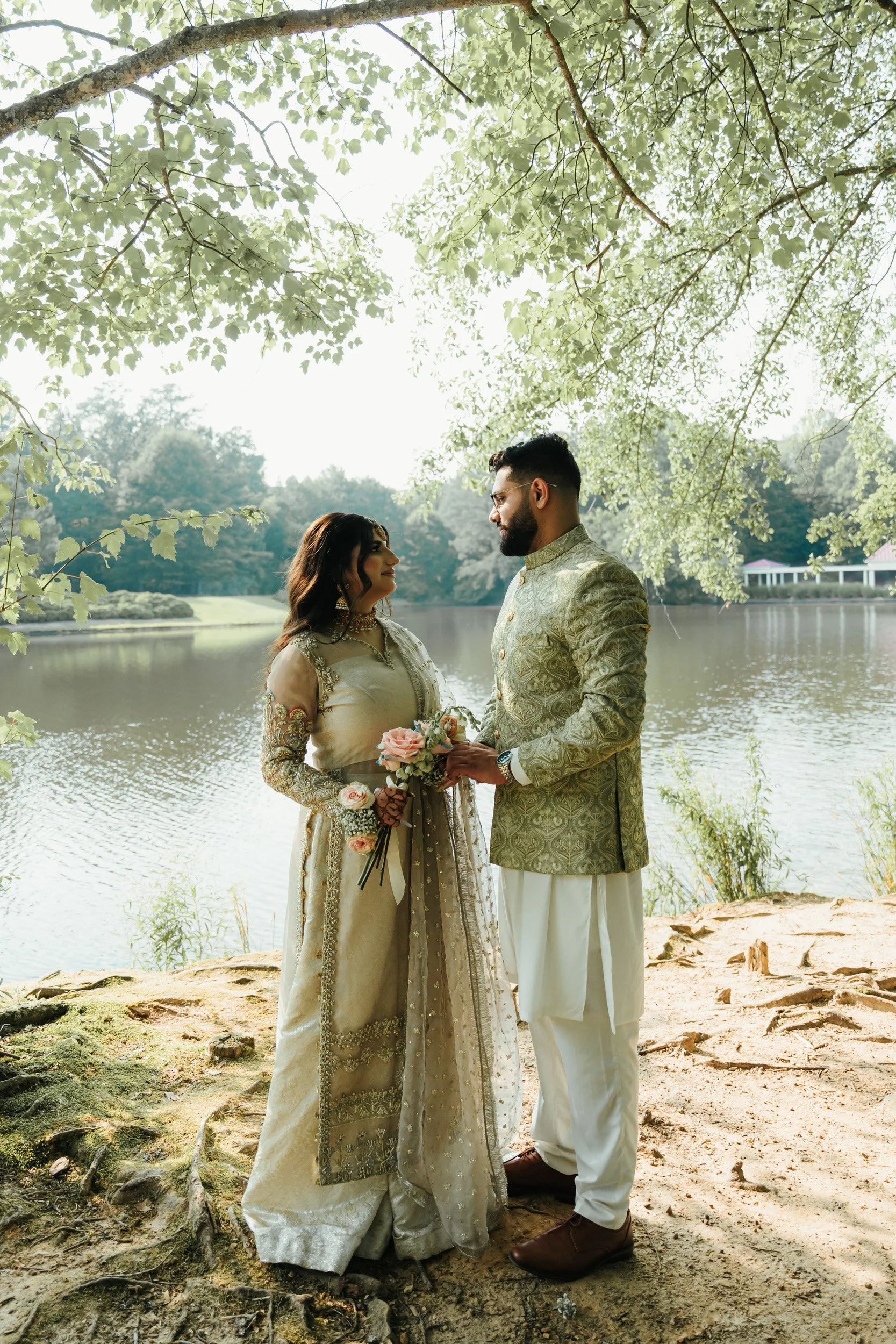 A couple dressed in traditional Indian wedding attire standing near a lake with trees overhead, holding a bouquet of pink roses.