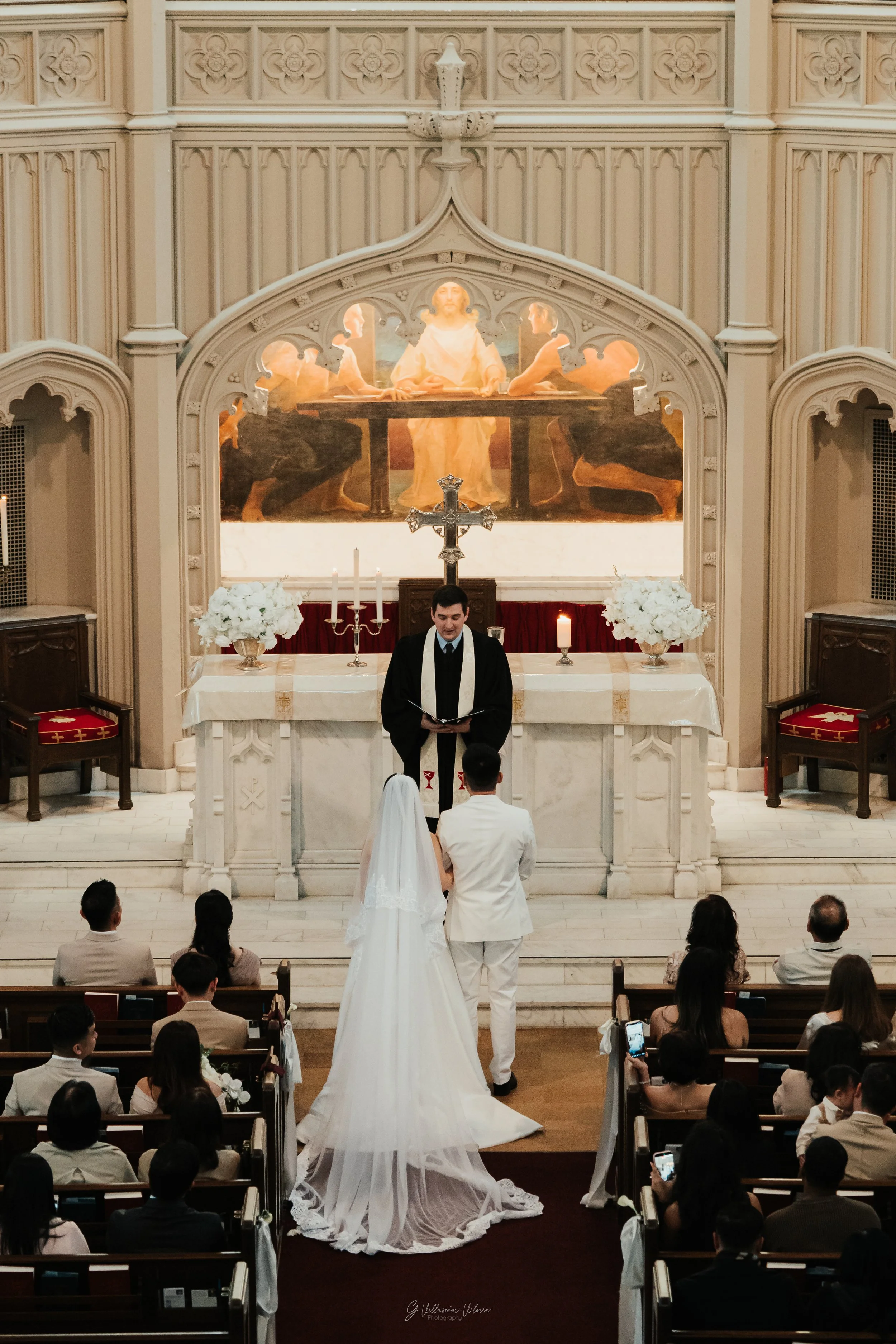 A wedding ceremony taking place inside a church with a bride in a long white gown and veil, and a groom in a white suit, standing before a priest at the altar. The altar is decorated with white flowers, candles, and a large cross, with a religious pa