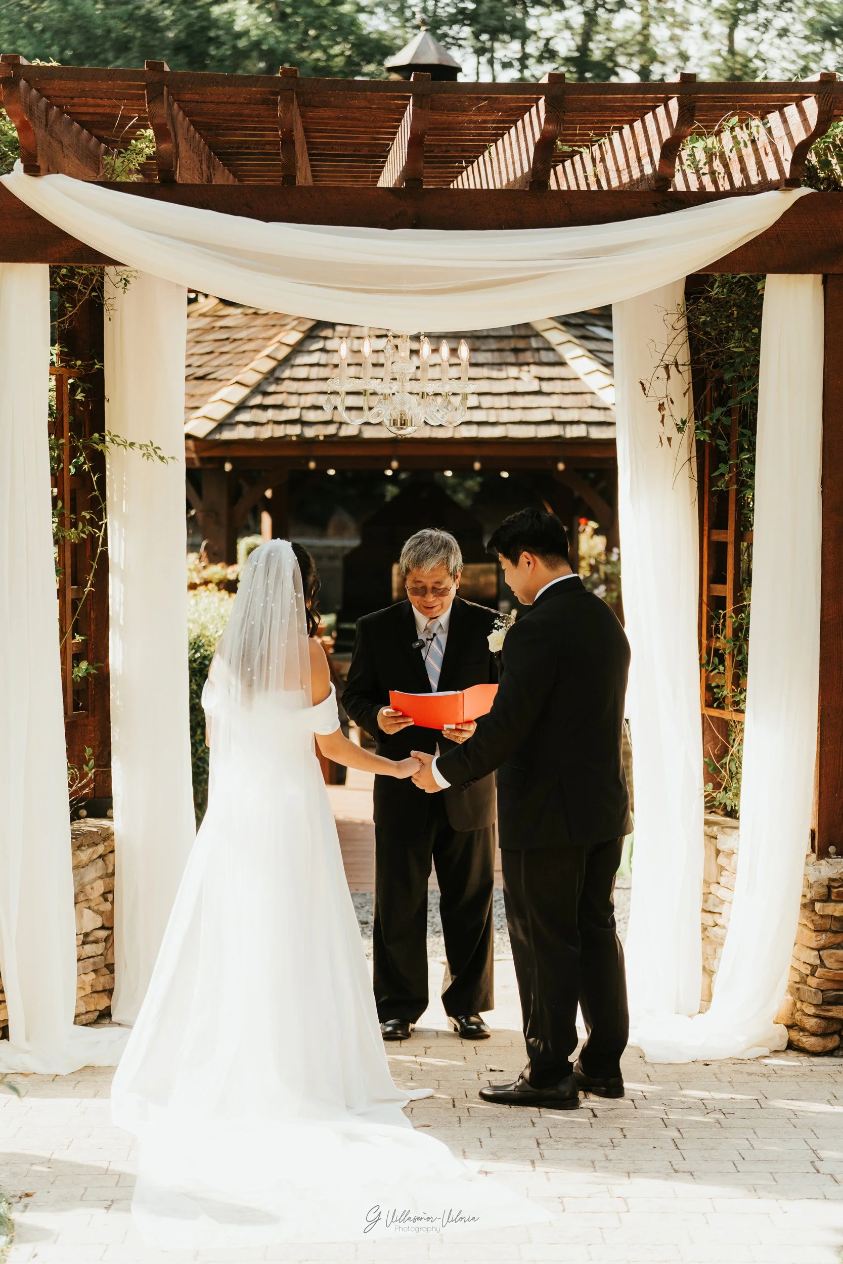 A wedding ceremony taking place outdoors under a wooden arch with white drapery, with a bride in a white gown and veil, a groom in a black suit, and a officiant standing in front of them, holding a red folder.