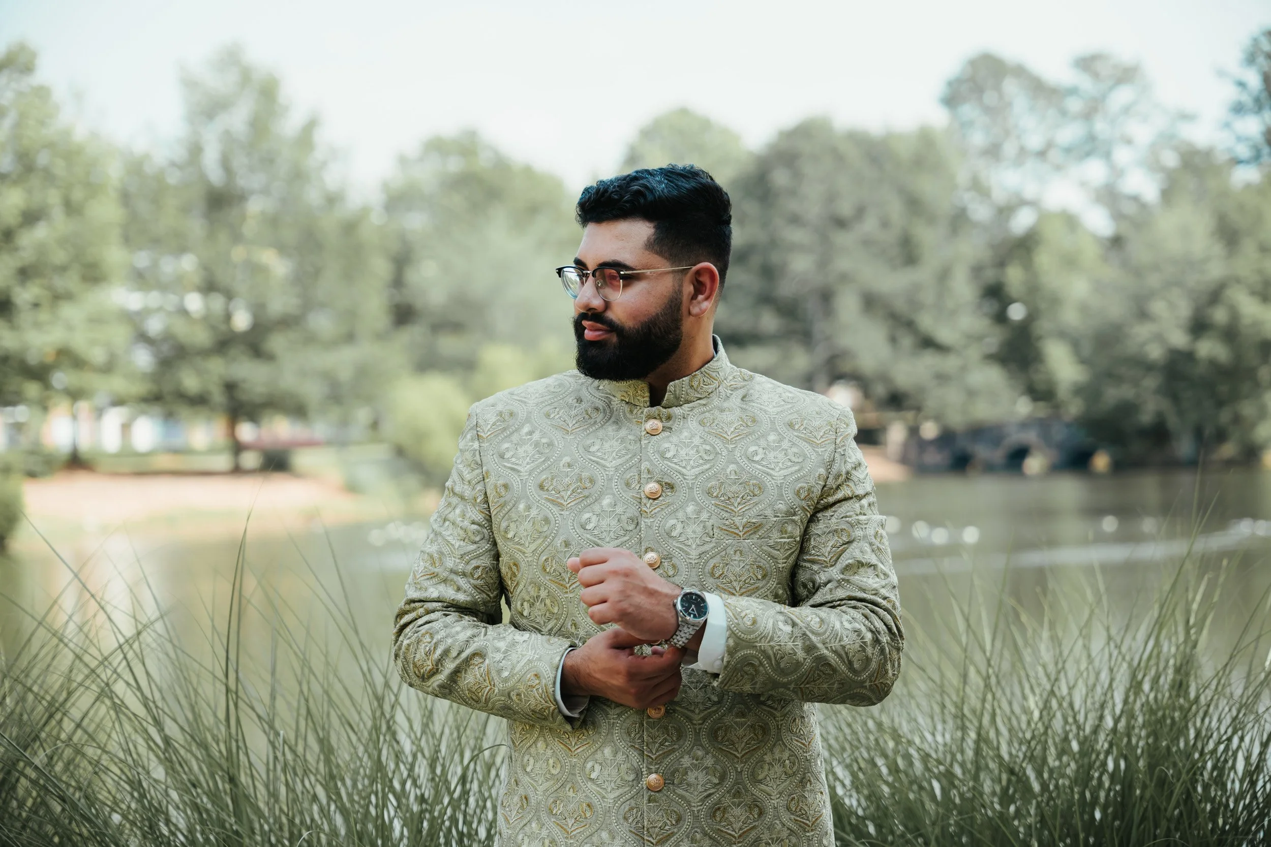 A man dressed in traditional Indian attire standing outdoors near a body of water with trees in the background.