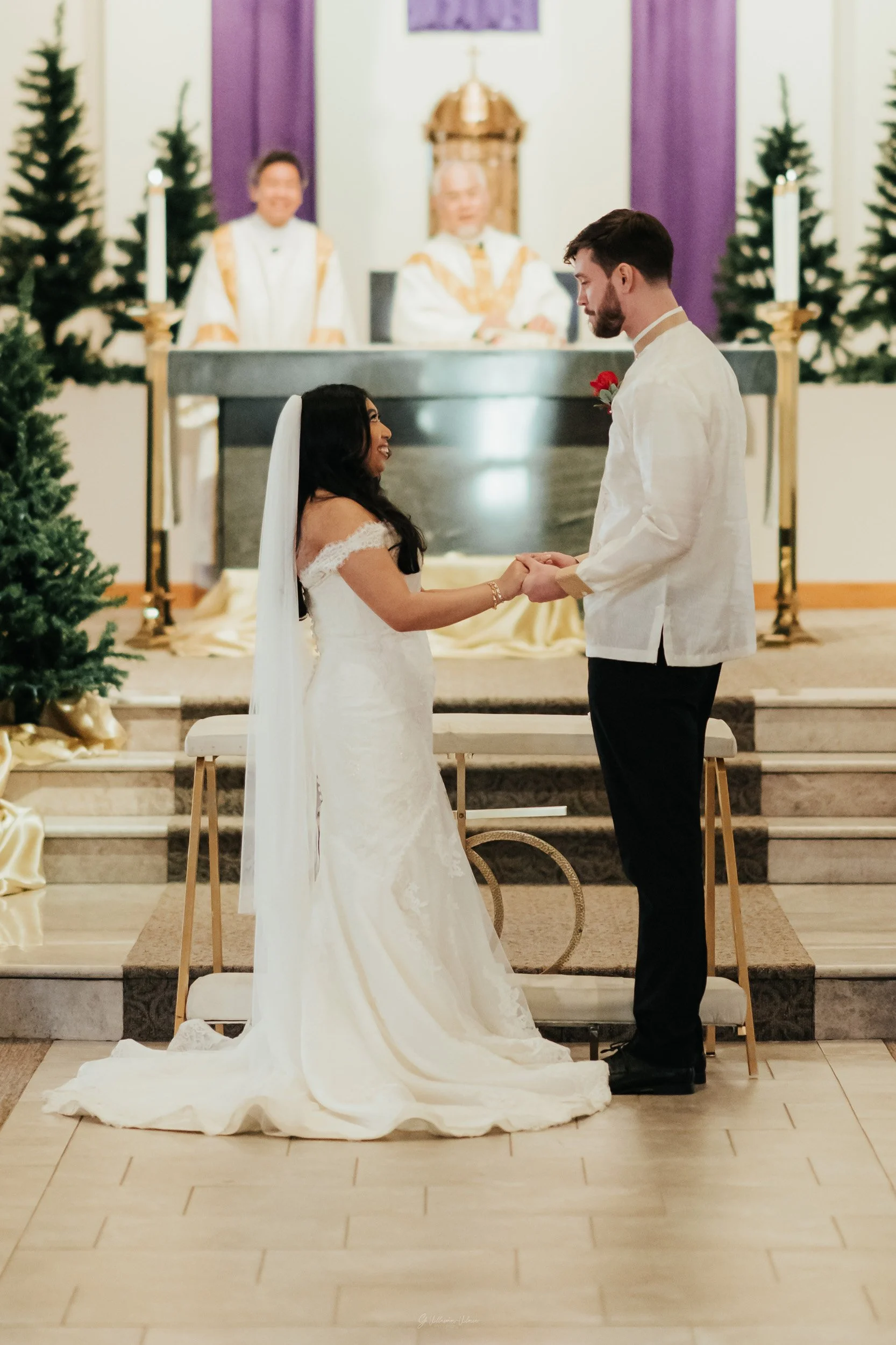 A bride and groom exchanging wedding vows during a ceremony in a church, with priests in the background and Christmas trees on either side.