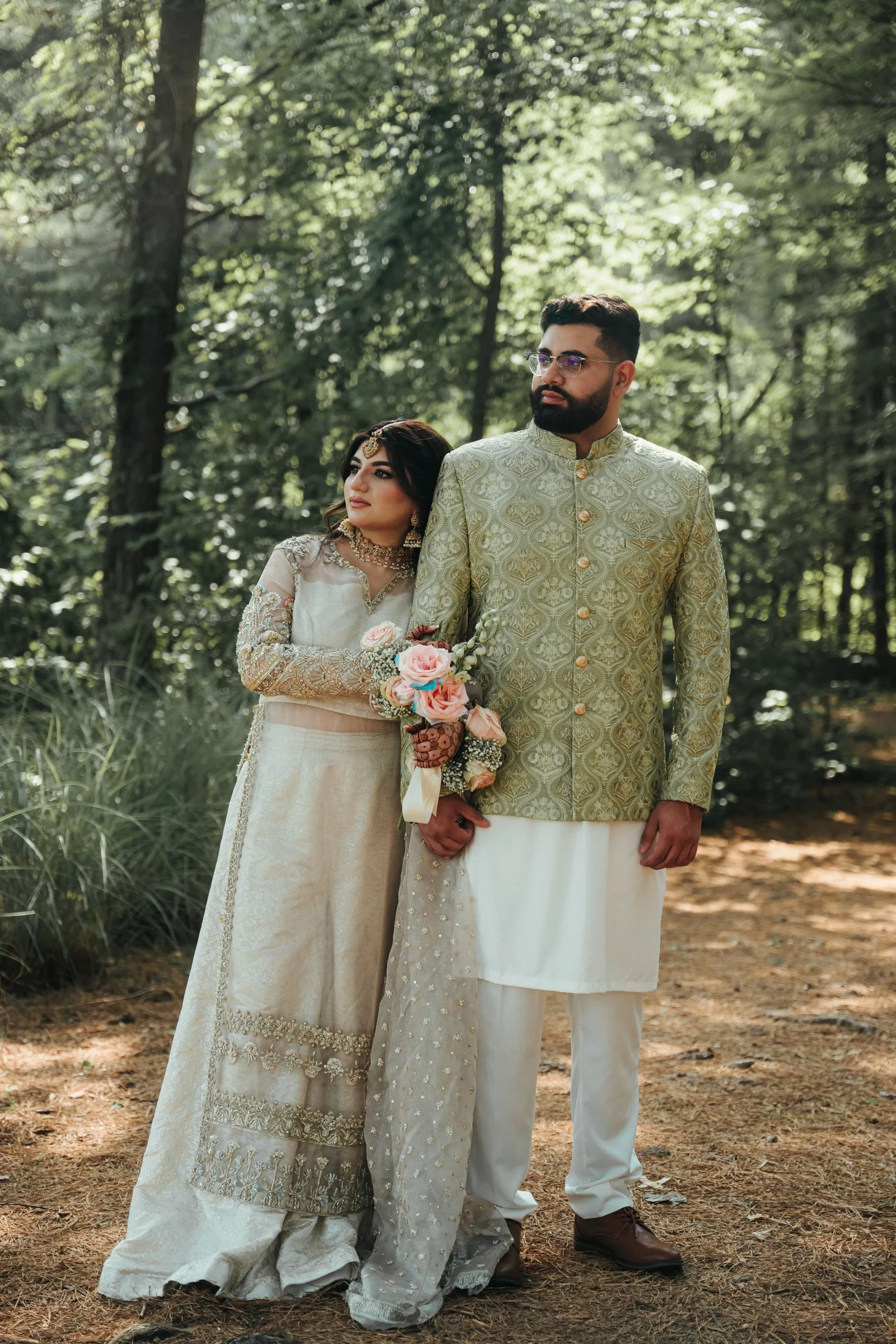 A couple dressed in traditional South Asian wedding attire standing in a wooded outdoor area, with the woman holding a bouquet of pink roses and the man wearing glasses.