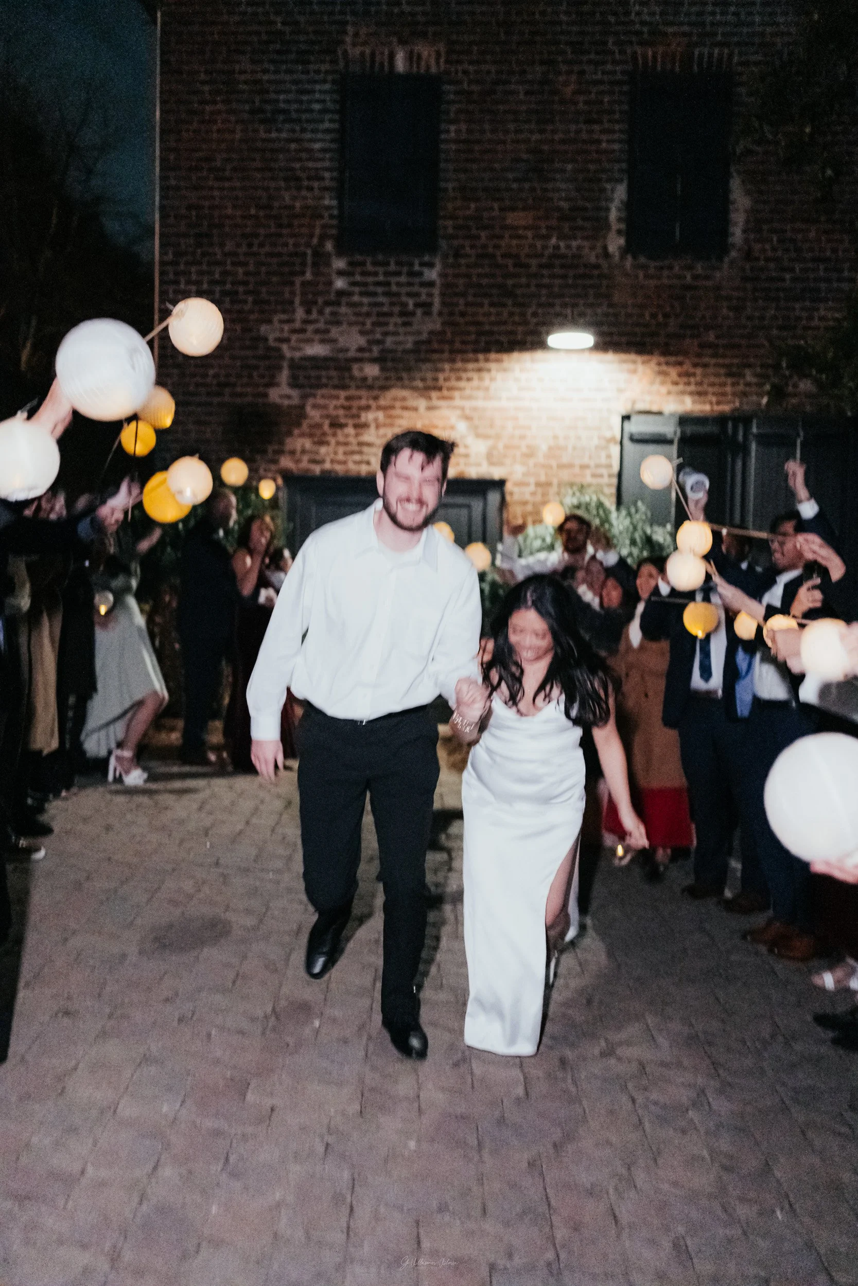 A couple is walking hand in hand at night, smiling, surrounded by a crowd holding paper lanterns on a cobblestone street.