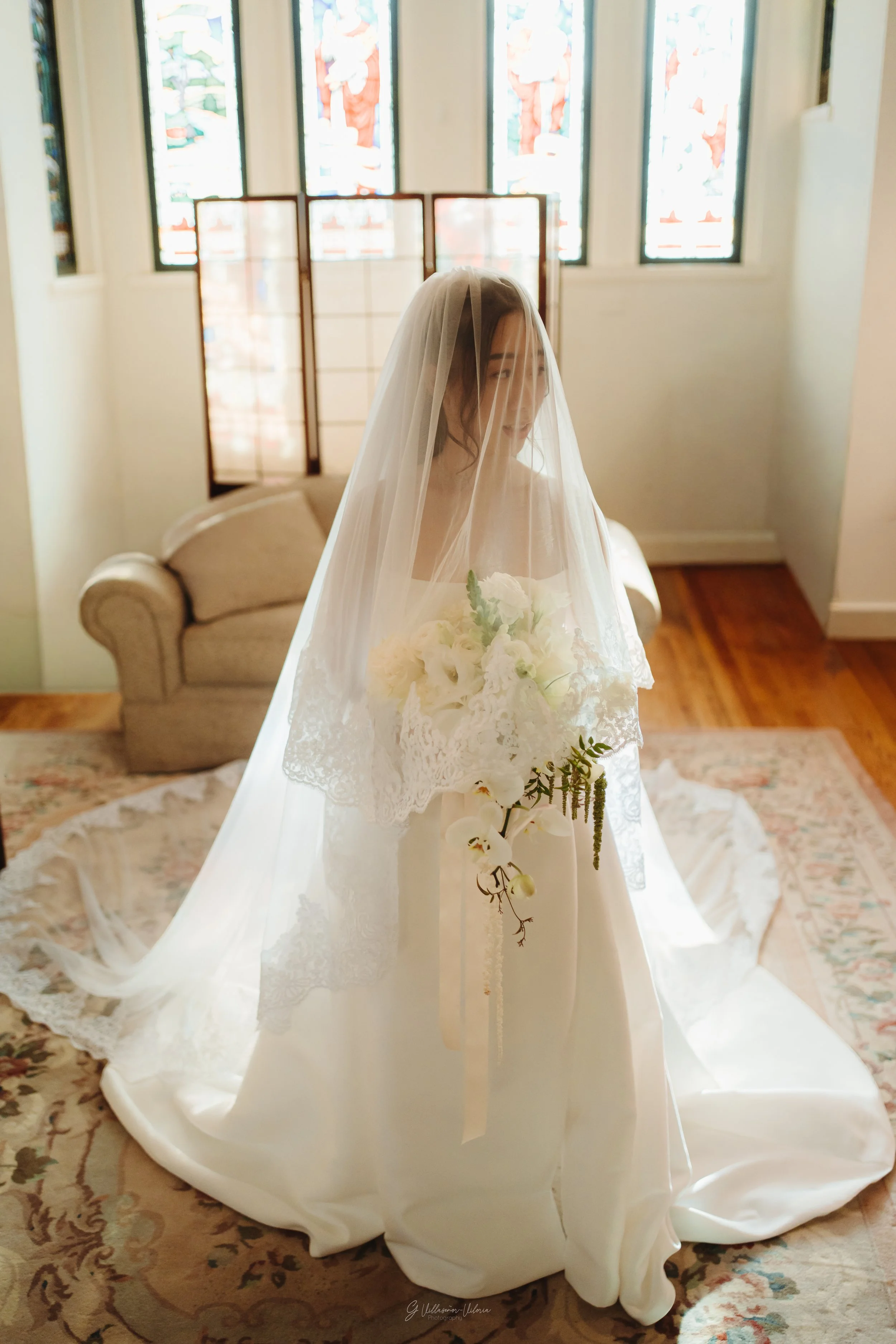 A bride in a white wedding gown with lace details, standing in a softly lit room with stained glass windows, holding a bouquet of white flowers, and wearing a veil.