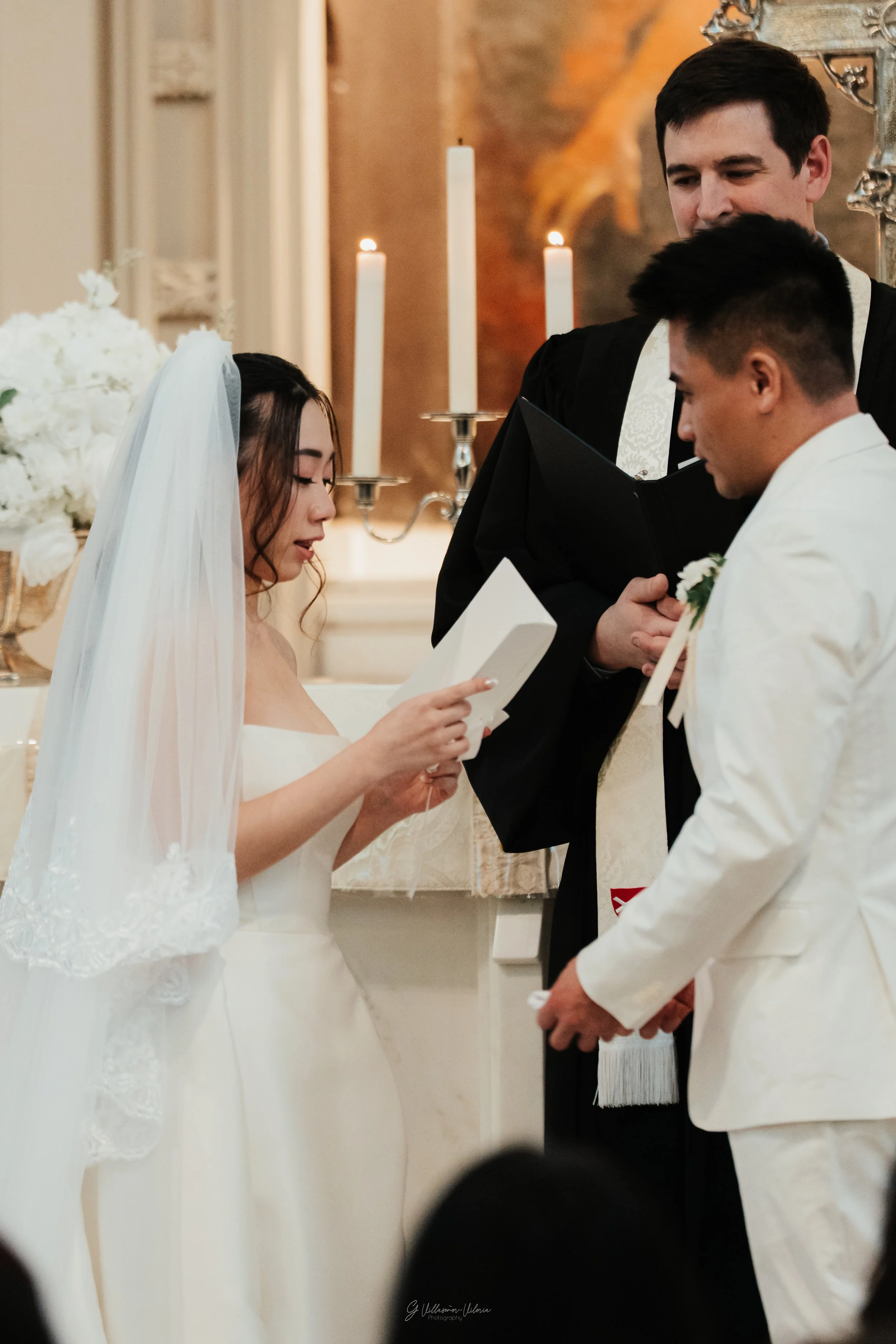 A bride and groom standing before an officiant during a wedding ceremony, with candles and flowers in the background.