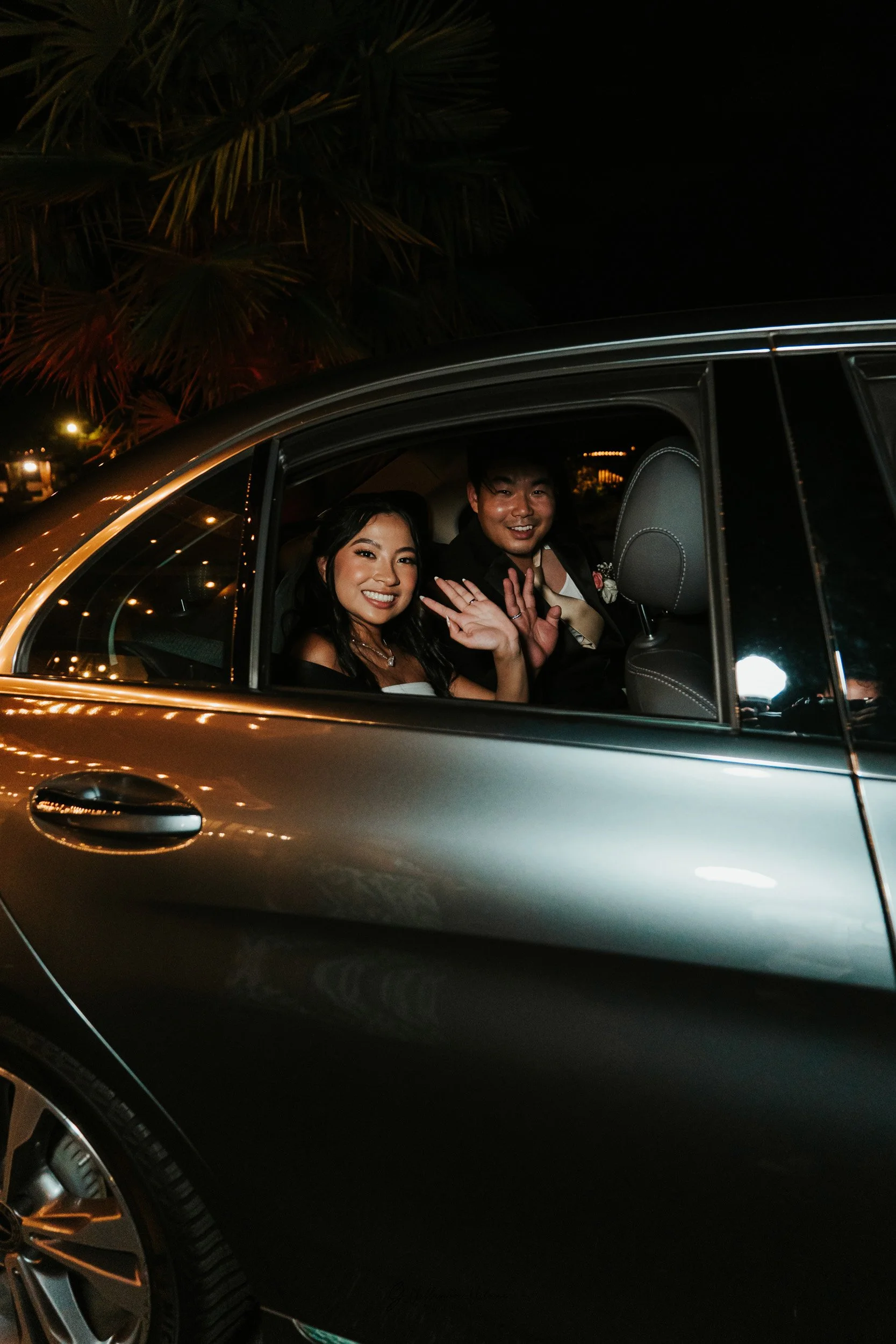 A smiling woman and man are sitting in a car at night, waving at the camera, with palm trees in the background.