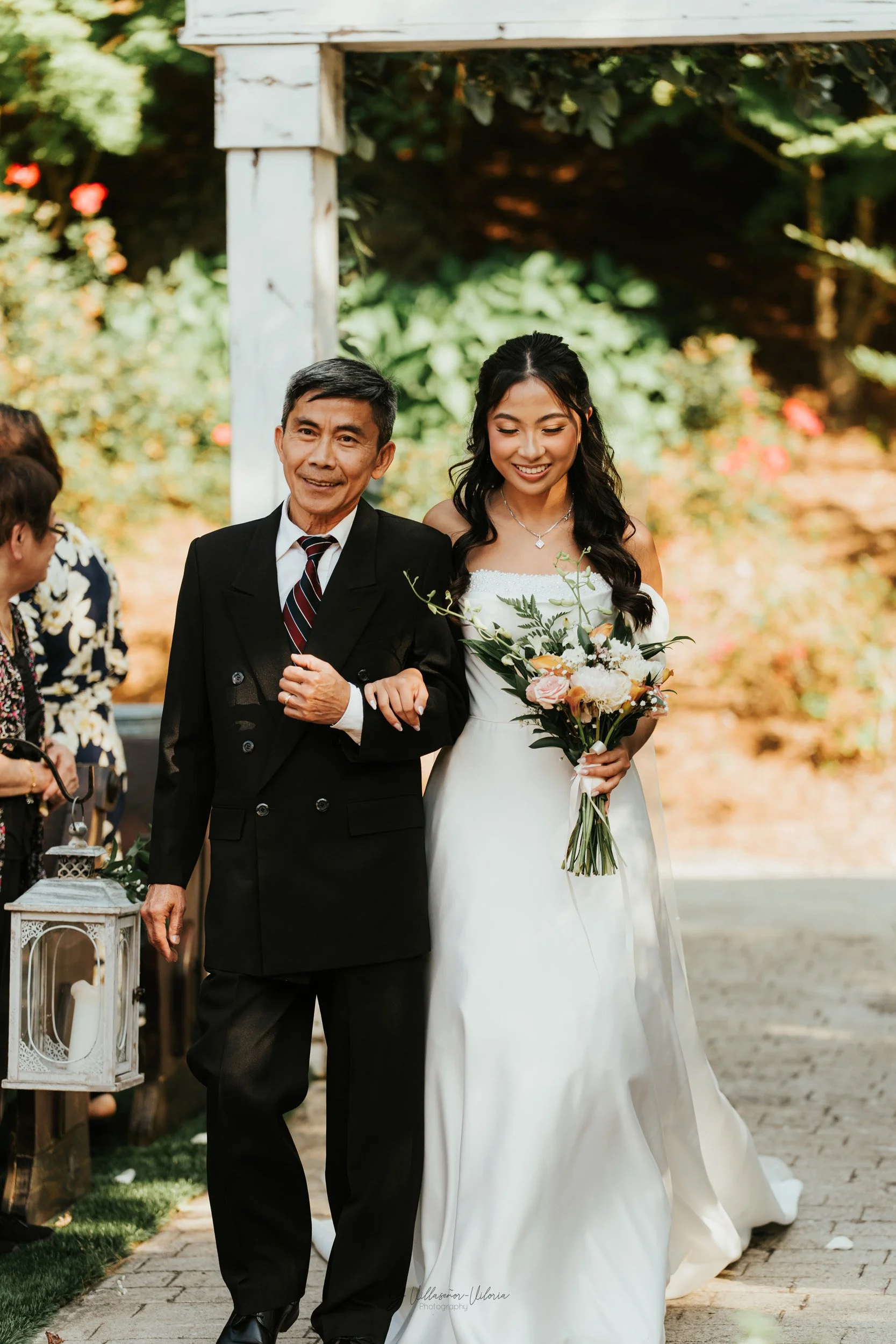 A bride in a white wedding dress holding a bouquet of flowers walking with an older man, likely her father, during a wedding ceremony outdoors.