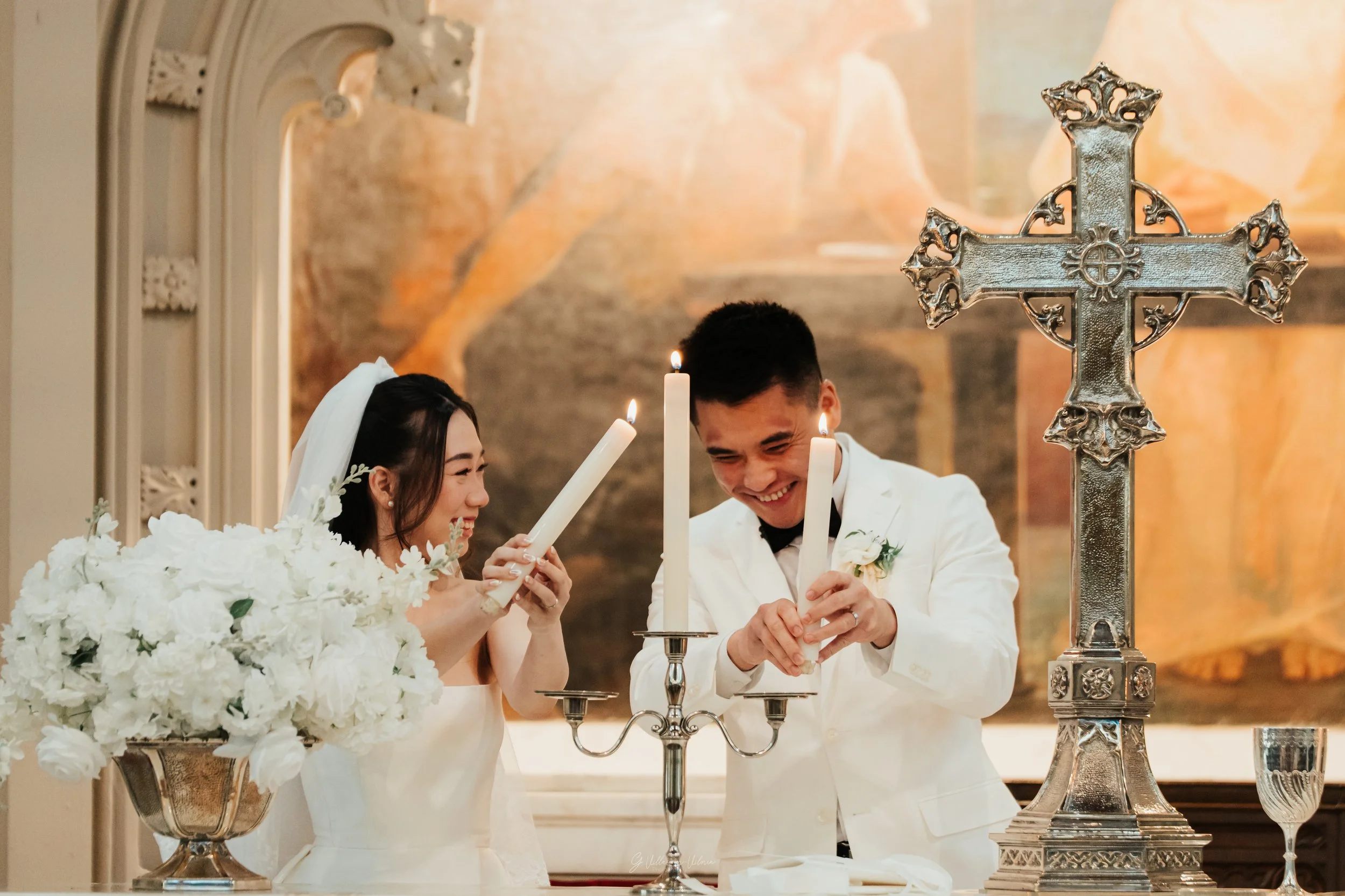 A bride and groom lighting candles during their wedding ceremony, with a decorative cross and floral arrangement on the altar.