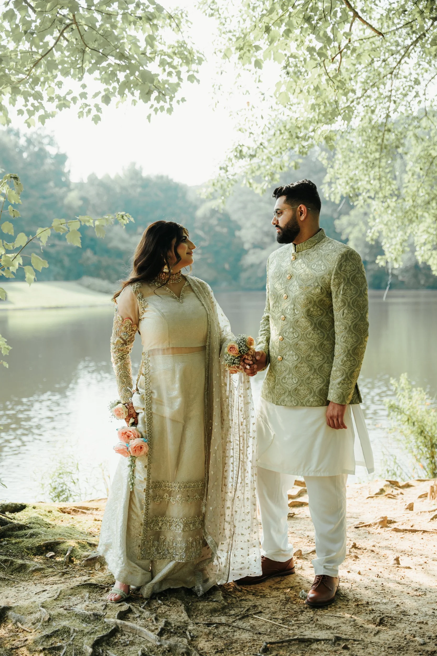 A couple dressed in traditional Indian wedding attire standing outdoors by a river, holding hands, with trees and greenery in the background.