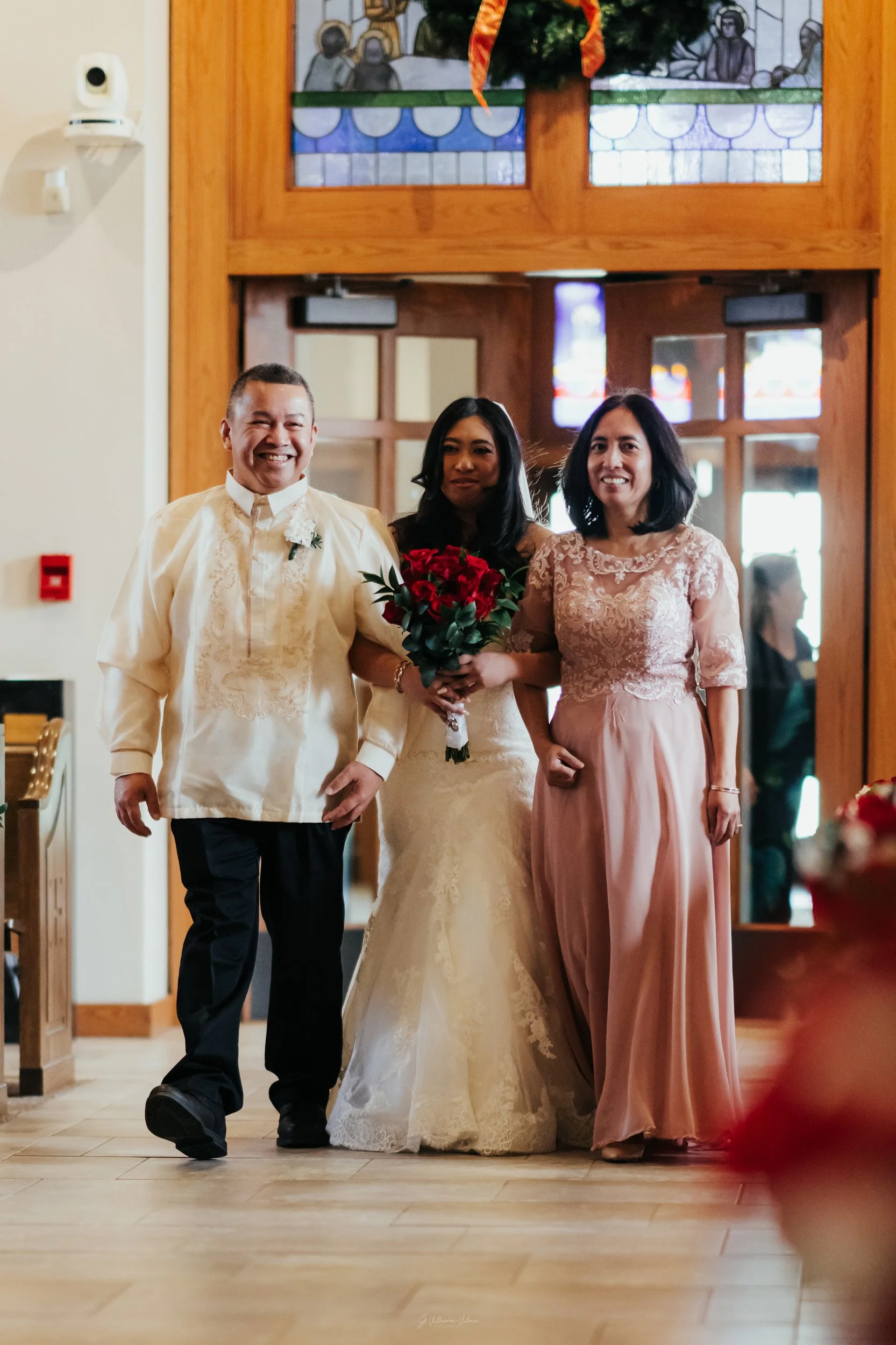 A bride in a lace wedding gown holding a bouquet of red roses, walking with a man in a cream barong and a woman in a pink lace dress inside a church.