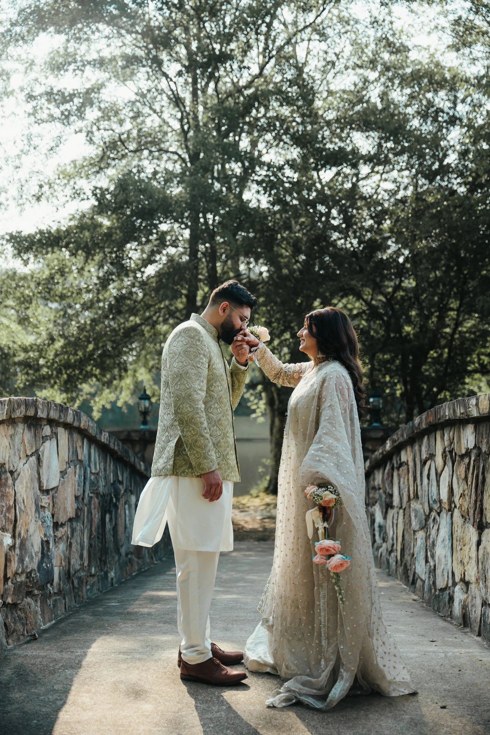 A bride and groom stand on a stone bridge, with trees in the background, as the bride feeds a cake slice to the groom during their wedding celebration.