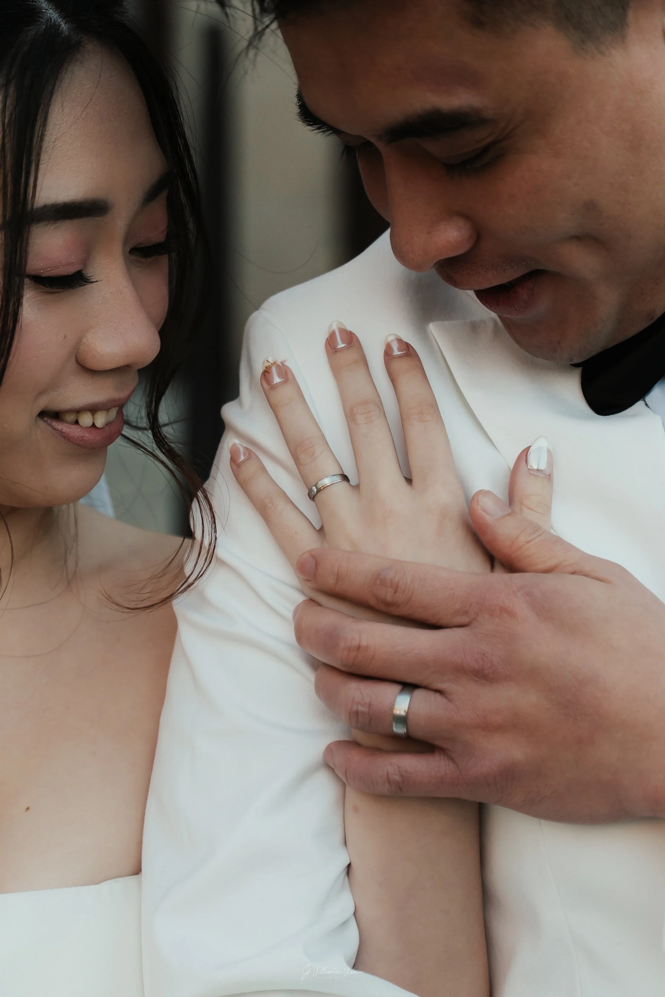 A close-up of a bride and groom sharing an intimate moment, showing the bride's face and hands and the groom's face and hands. They are wearing wedding rings and the groom is in a white tuxedo.