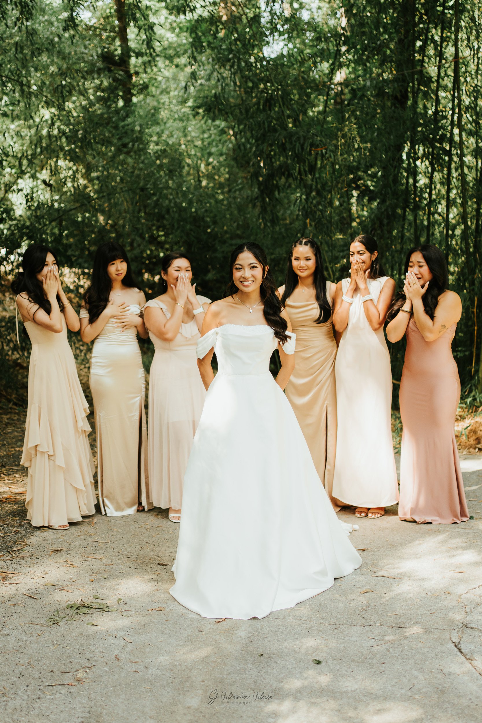 A bride in a white wedding dress smiling in front of six women in pastel dresses, all outdoors with green trees in the background.