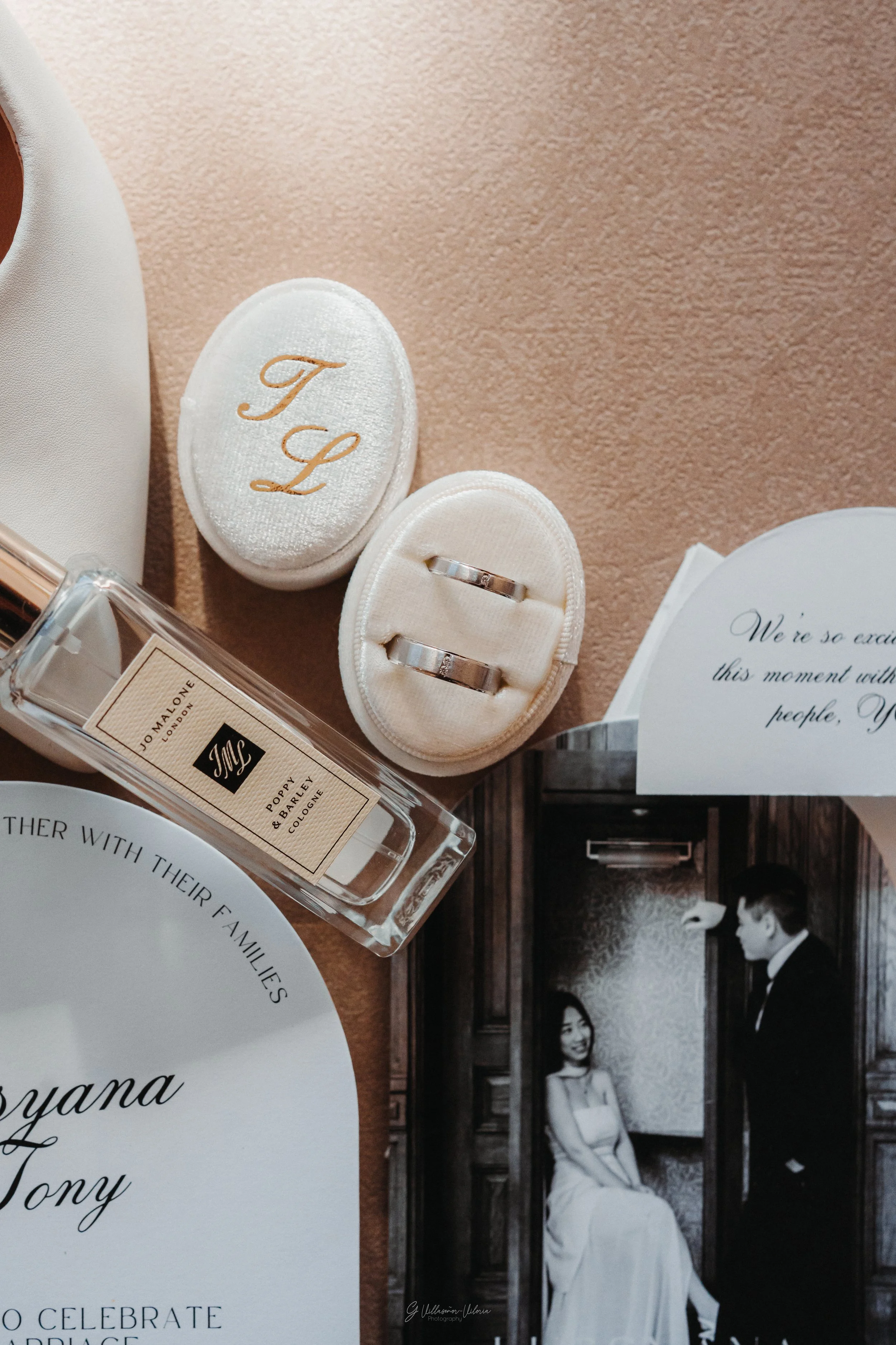 Wedding rings in a white velvet ring box, a perfume bottle labeled 'Jo Malone London Poppy & Barley Cologne,' a black and white photo of a bride and groom, and wedding invitations with elegant script on a table.