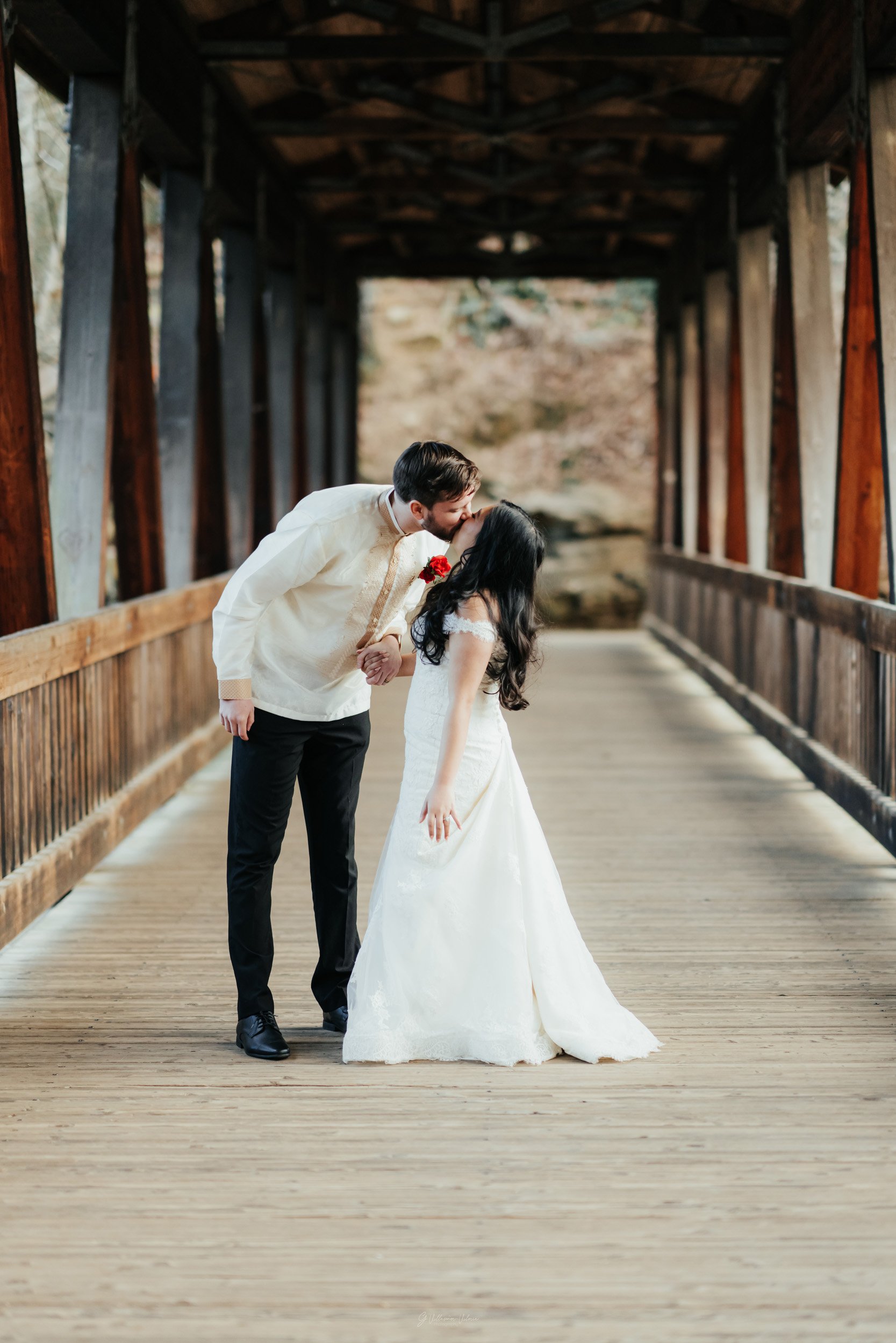 Couple in wedding attire sharing a kiss inside a wooden covered bridge.
