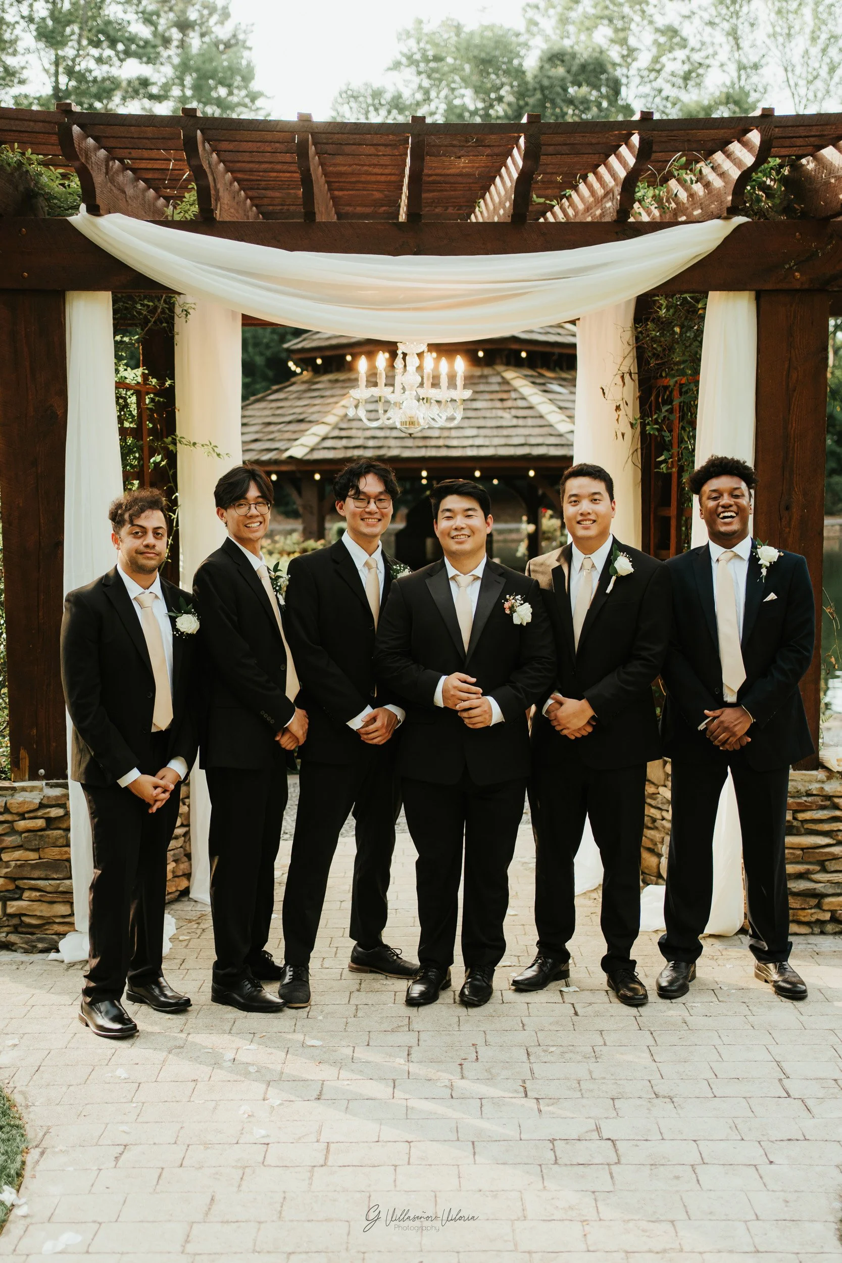 A group of six men in suits standing under a wedding arch with drapery, on a stone patio outside, with trees and a wooden structure in the background.