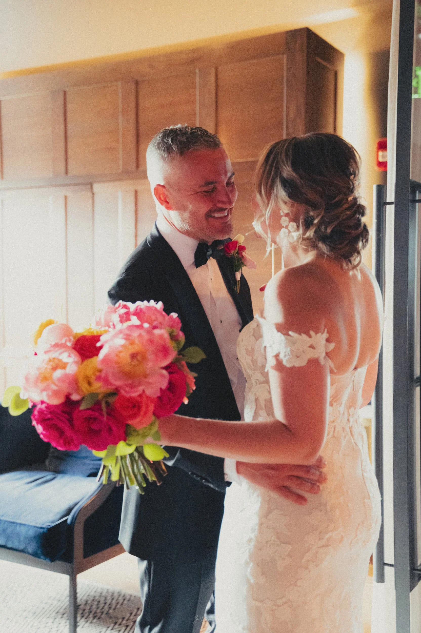 A bride and groom sharing a moment indoors, with the bride holding a colorful bouquet, and both smiling.