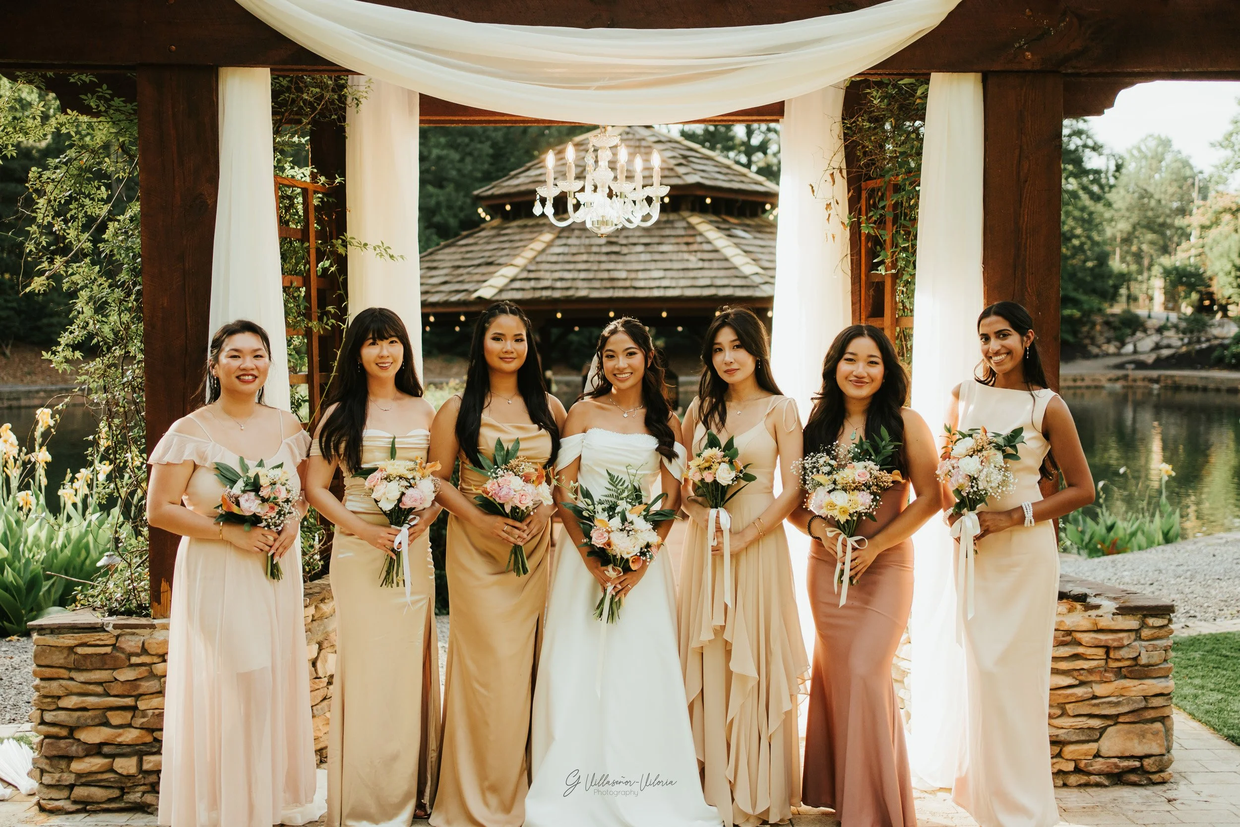 A group of eight women dressed in elegant gowns holding bouquets, standing under a decorated outdoor structure with drapes and a chandelier, near a pond or lake.