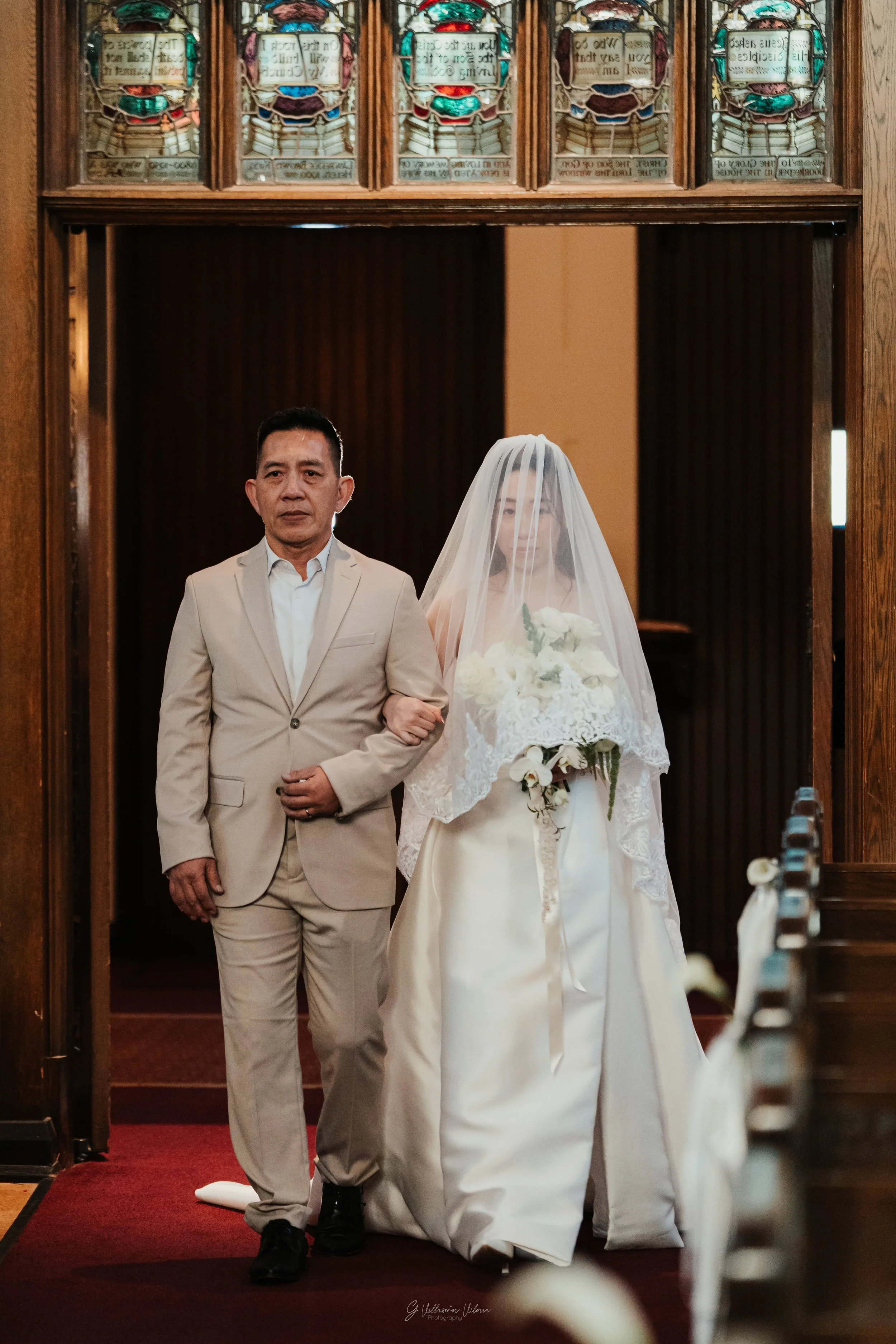 A bride in a white wedding gown and veil holding a bouquet of white flowers walking down the aisle with her father, inside a church with wooden accents and stained glass windows.