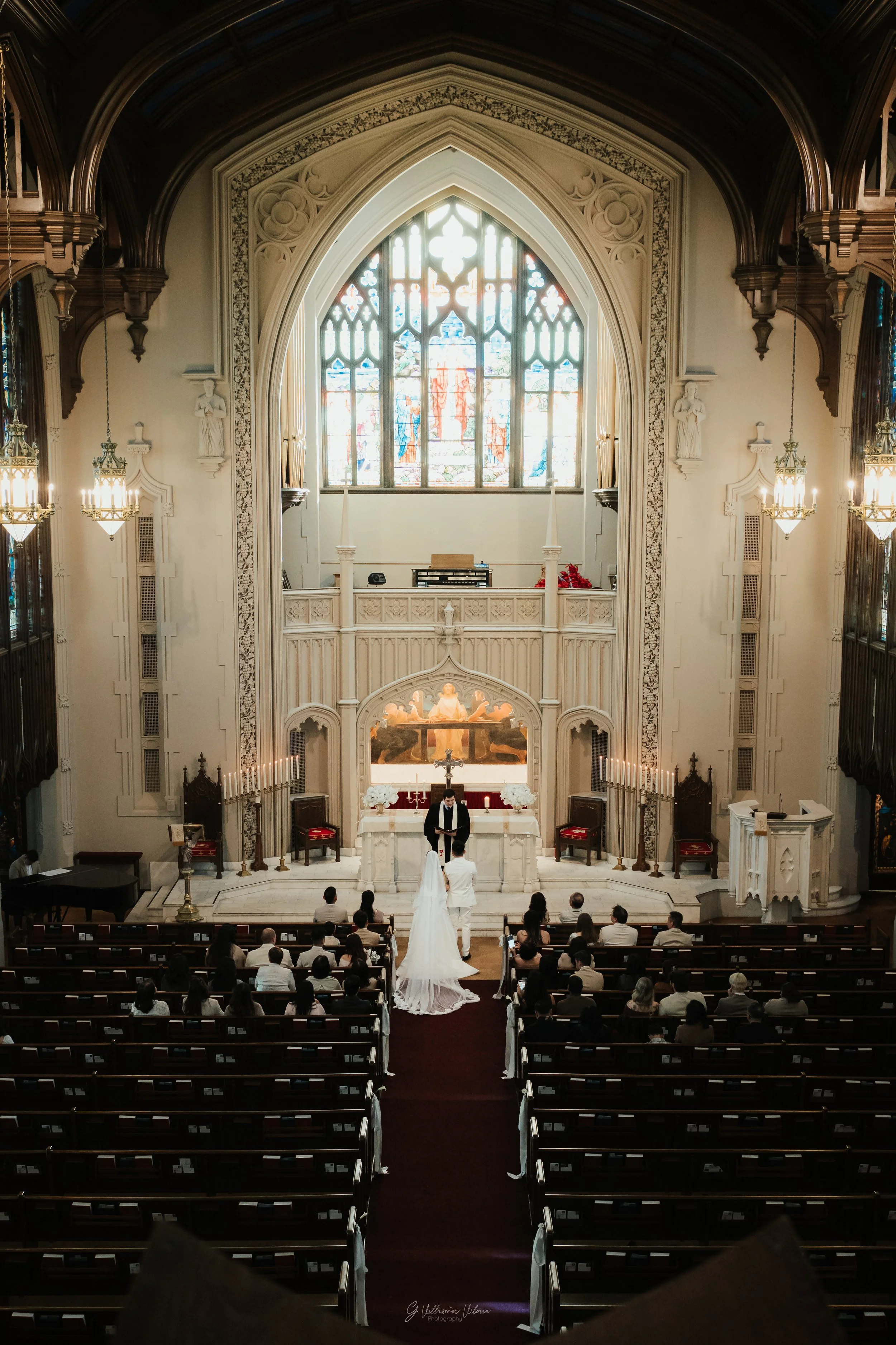 A wedding ceremony taking place inside a church with high ceilings, stained glass windows, and ornate architecture. The bride and groom are standing at the altar facing each other, with a priest officiating. Guests are seated in pews watching the cer
