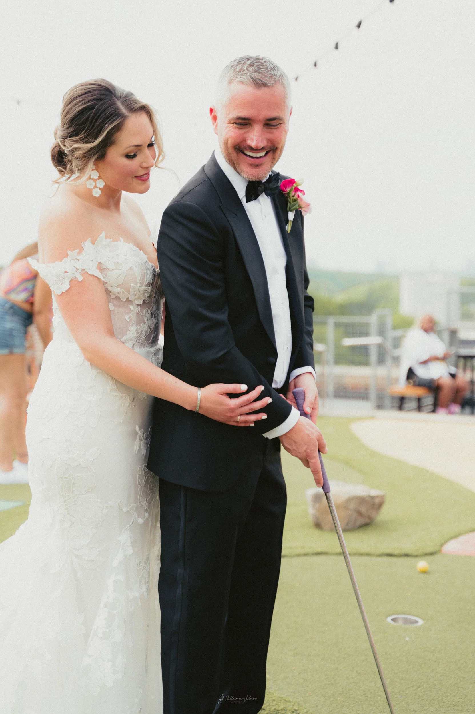 A bride and groom playing mini golf outdoors during their wedding celebration, with the groom holding a putter and smiling.