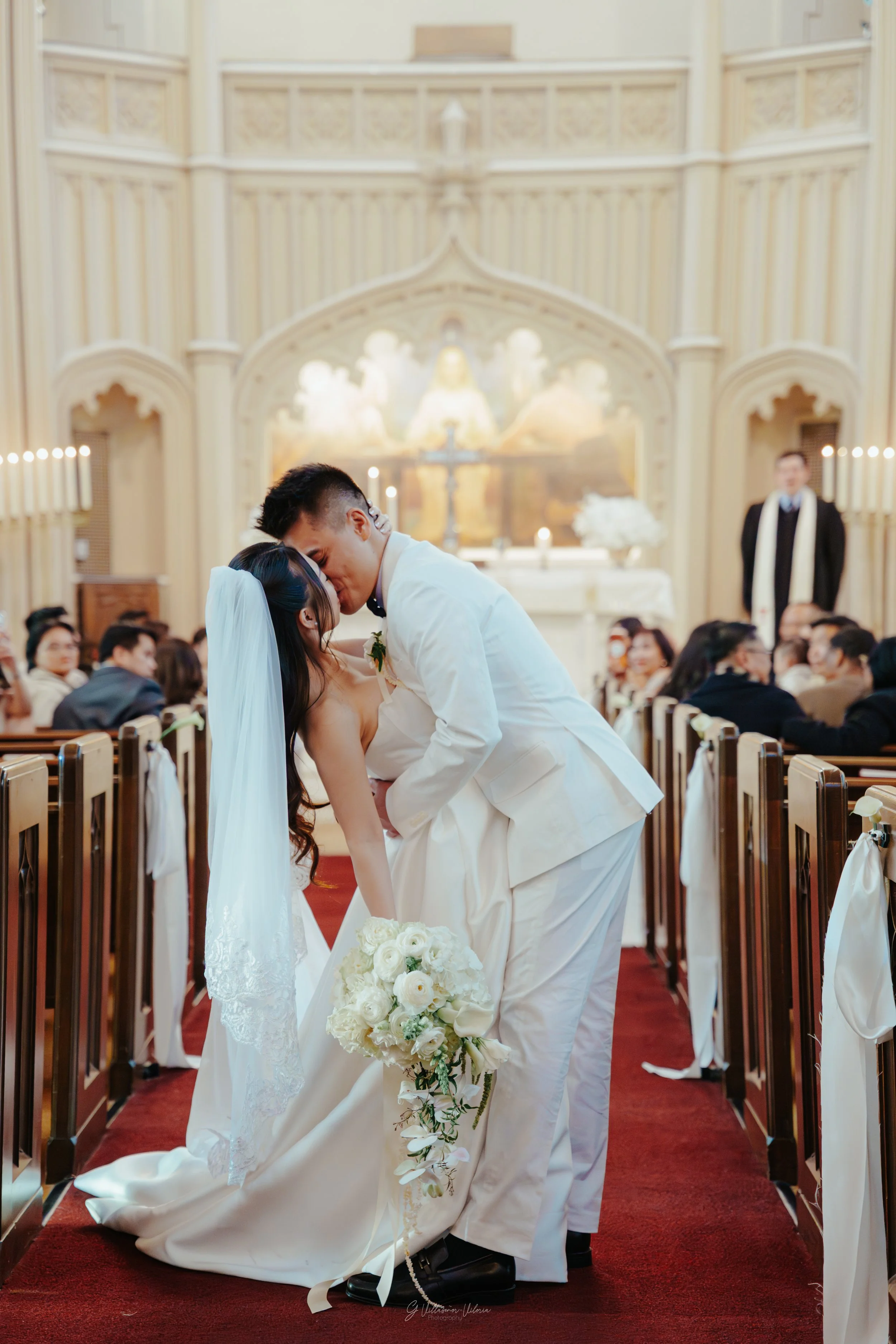 A bride and groom share a kiss during their wedding ceremony inside a church, surrounded by seated guests and lit by candles.