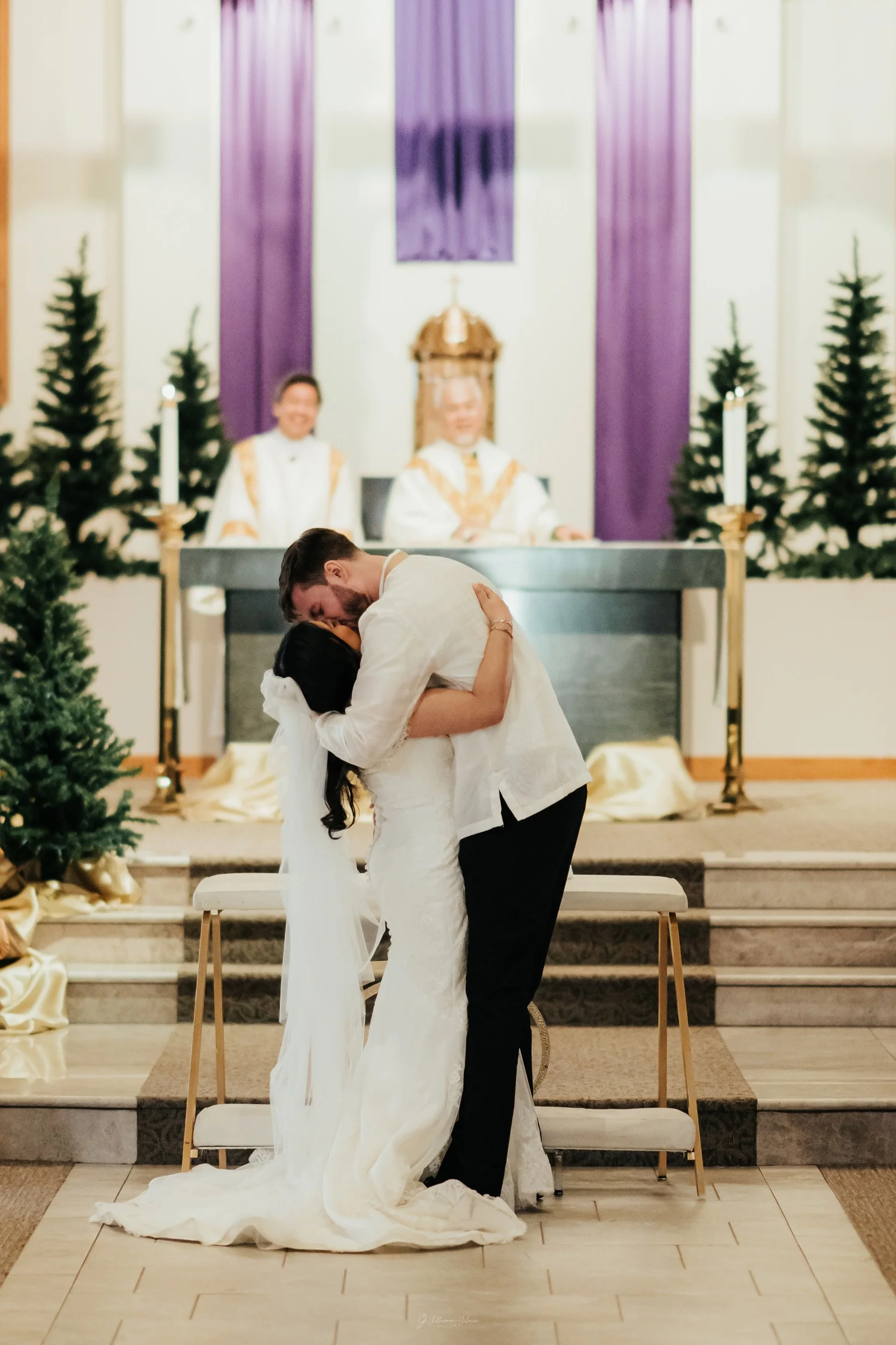 A bride and groom share a kiss during their wedding ceremony inside a church decorated with Christmas trees and gold ribbons.