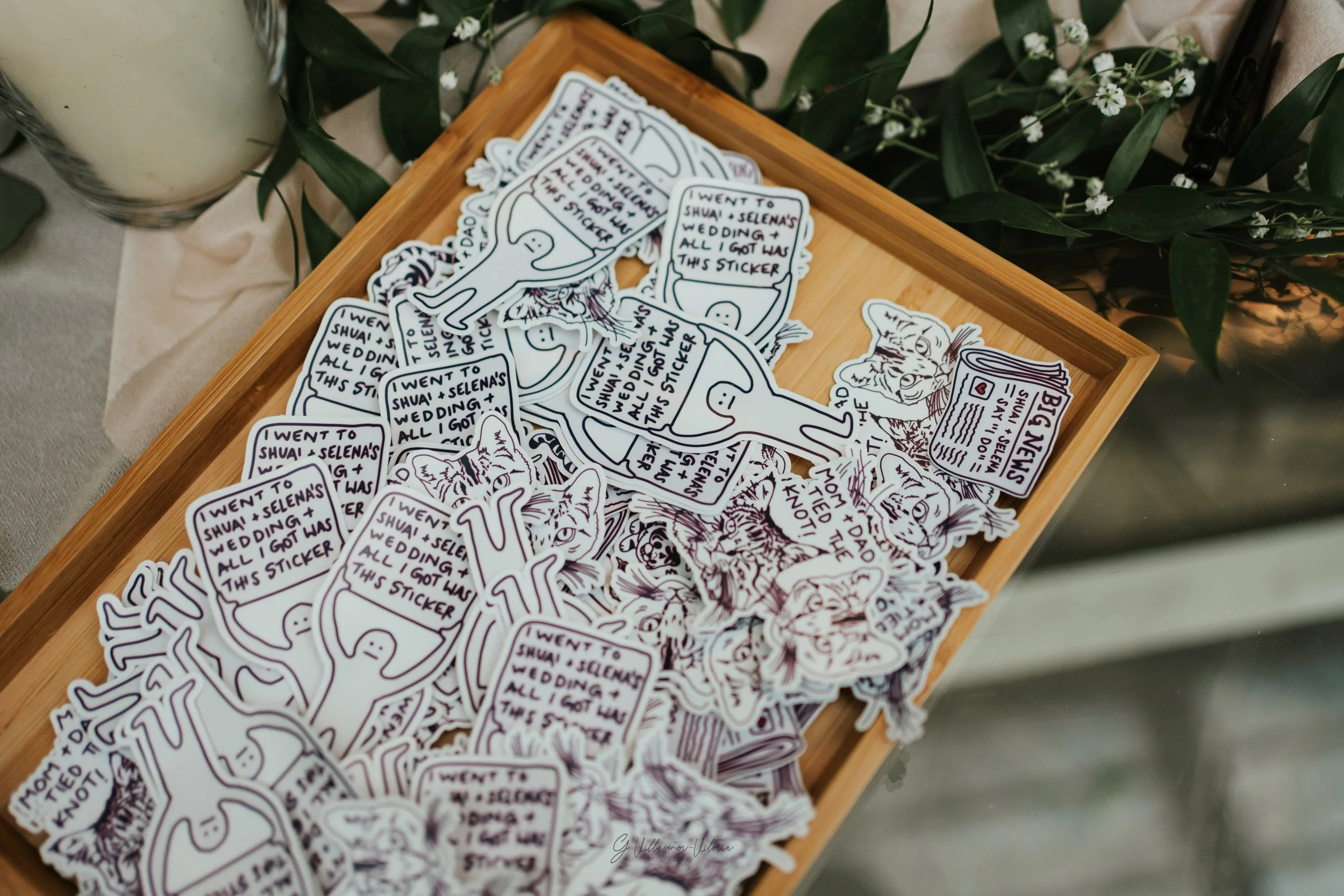 A wooden tray filled with various stickers featuring illustrations of cats, dinosaurs, and speech bubbles with texts like "I went to shua + Selena's wedding + all I got was this sticker." There is greenery adjacent to the tray and a candle on the lef